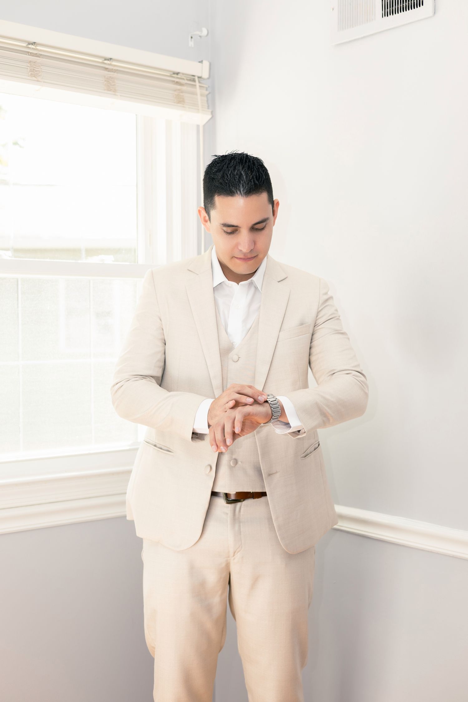 A person in a cream-colored suit checks their watch while standing in a bright white room with natural lighting.