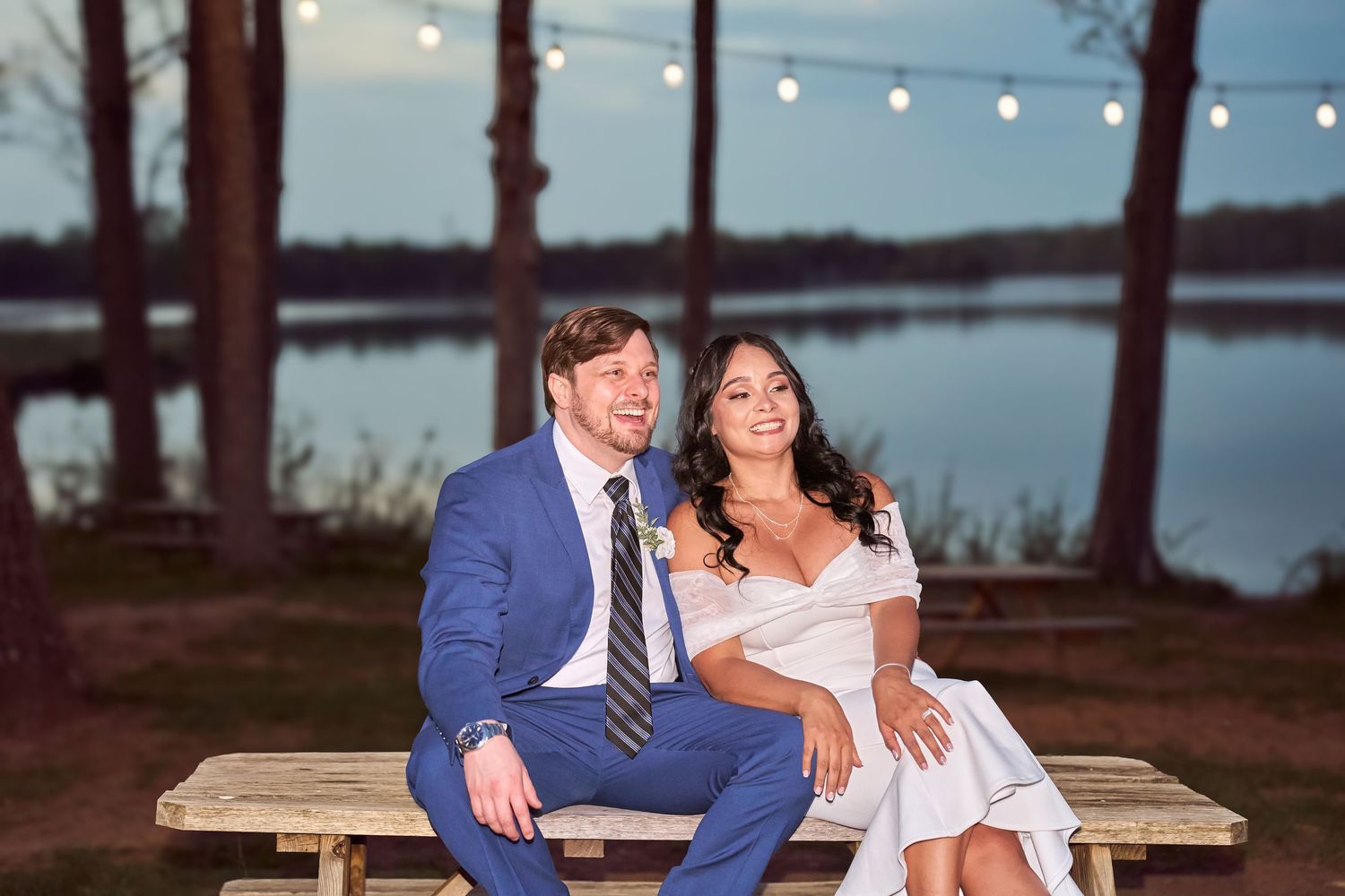 Bride and groom sitting on a wooden bench by a lake at dusk with string lights glowing behind them.