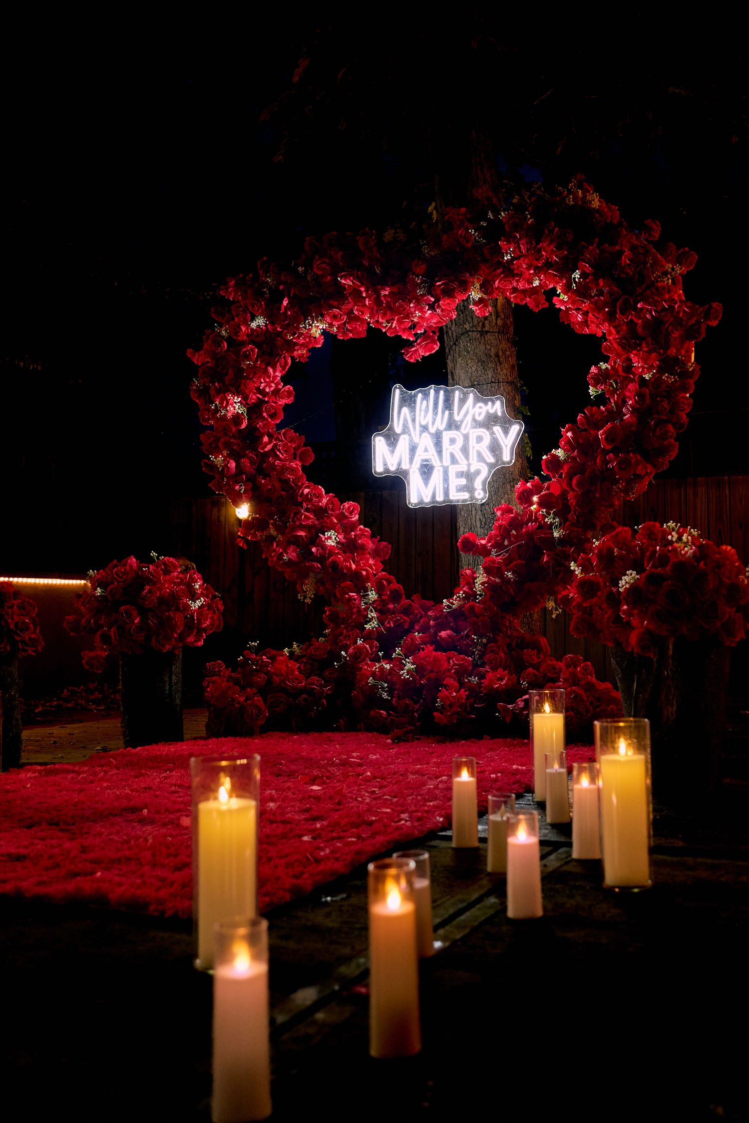 A romantic proposal setup with glowing candles leading to an illuminated 'Marry Me' sign surrounded by red floral decor.