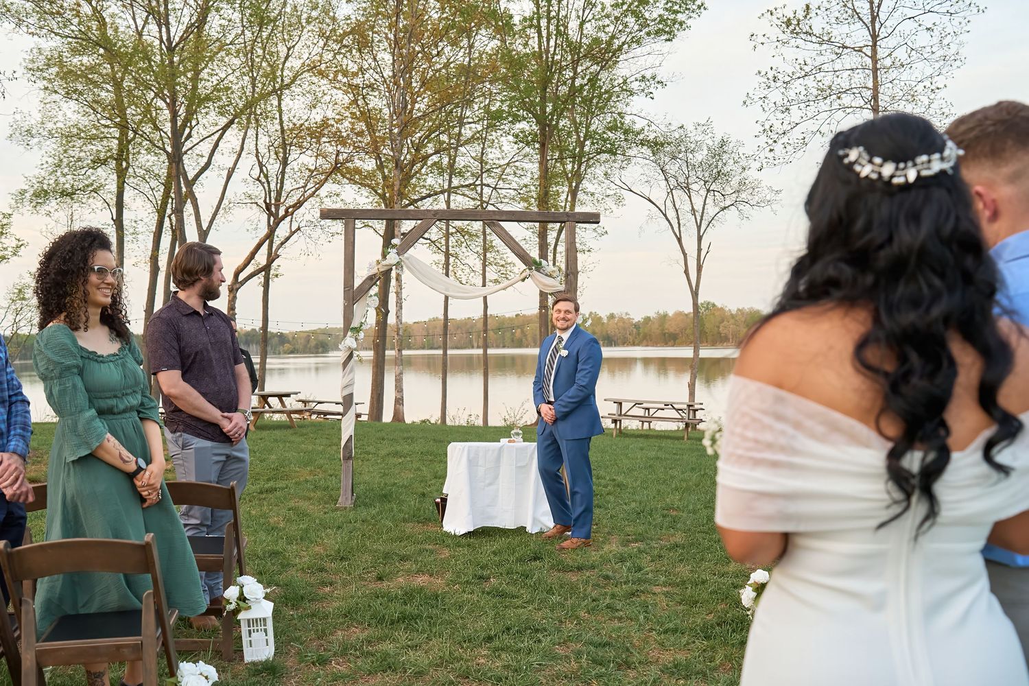 Outdoor wedding ceremony by a lake with groom in blue suit awaiting bride in white dress and floral crown.