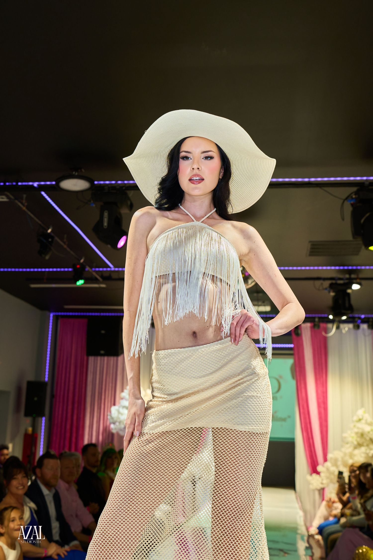Model walks runway in white fringed crop top, mesh skirt and wide-brim hat at fashion show.