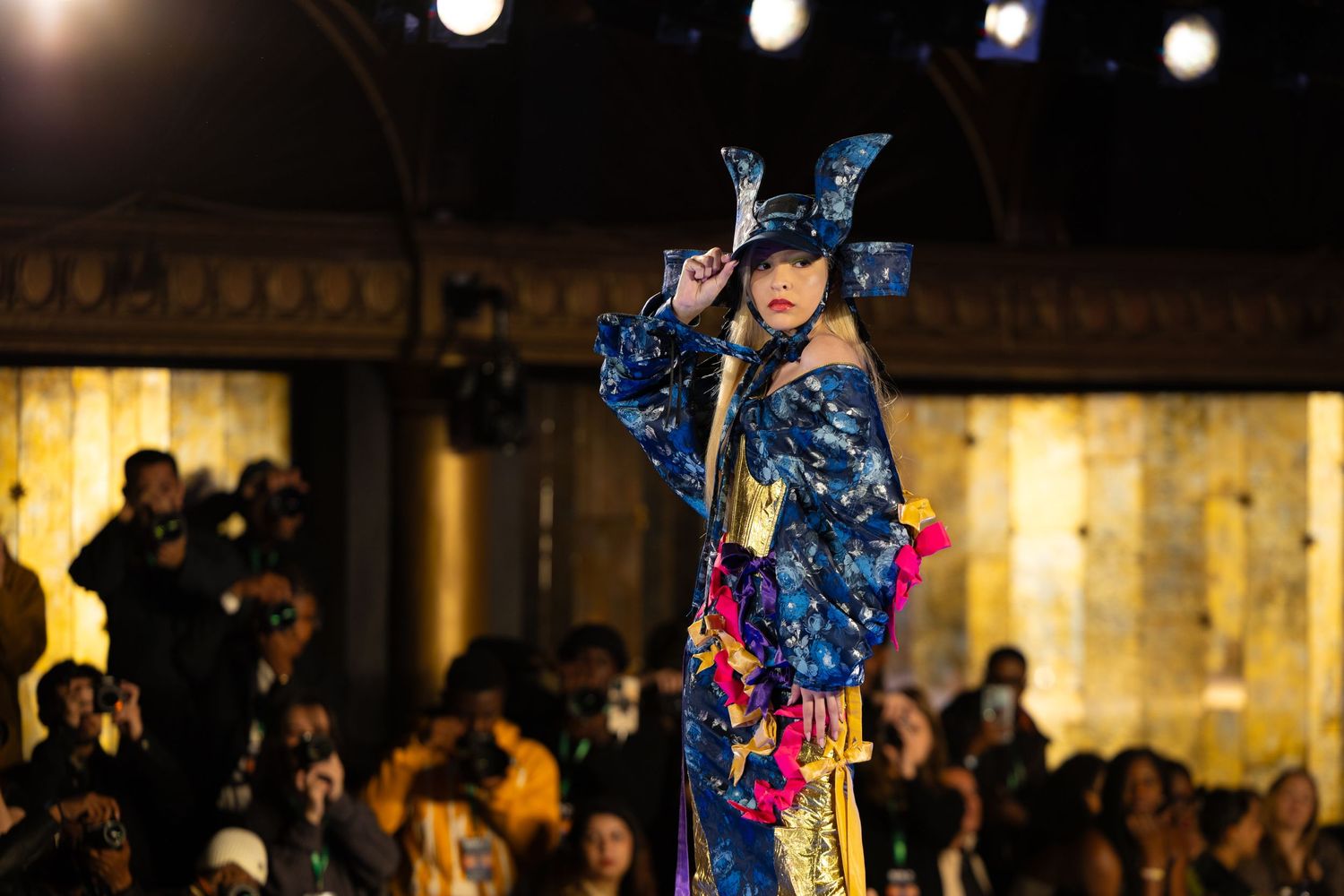Model in vibrant blue butterfly-inspired costume strikes pose on illuminated fashion runway.