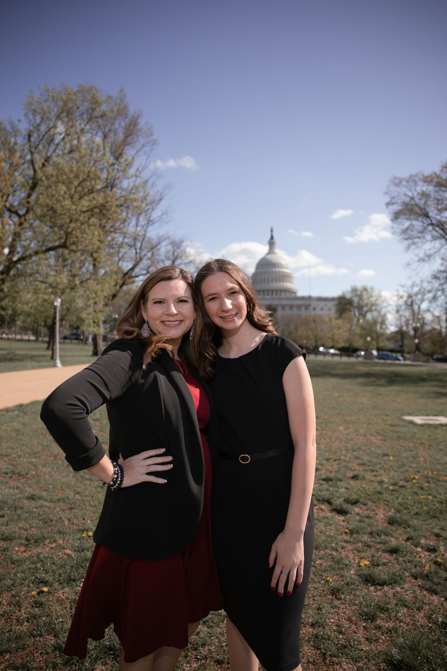 Two people posing in black dresses with the US Capitol dome visible in the background on a sunny day.
