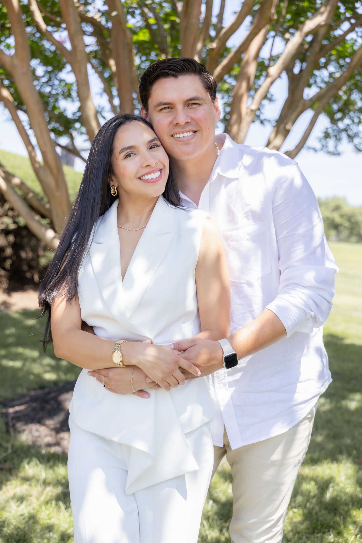 A couple in white outfits embraces while standing outdoors under a sunlit tree.