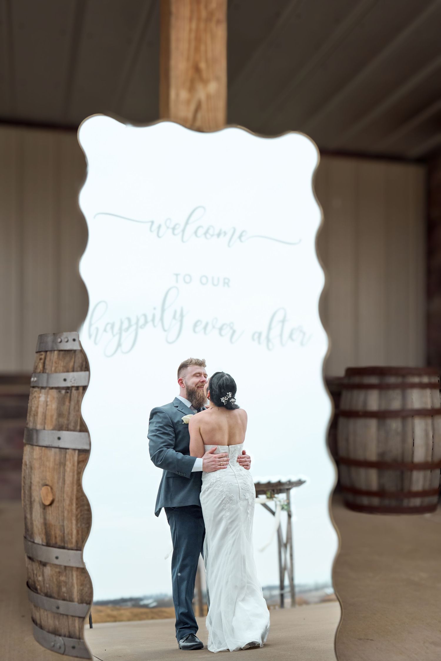 A couple embraces in front of a rustic welcome sign framed by wooden barrels.