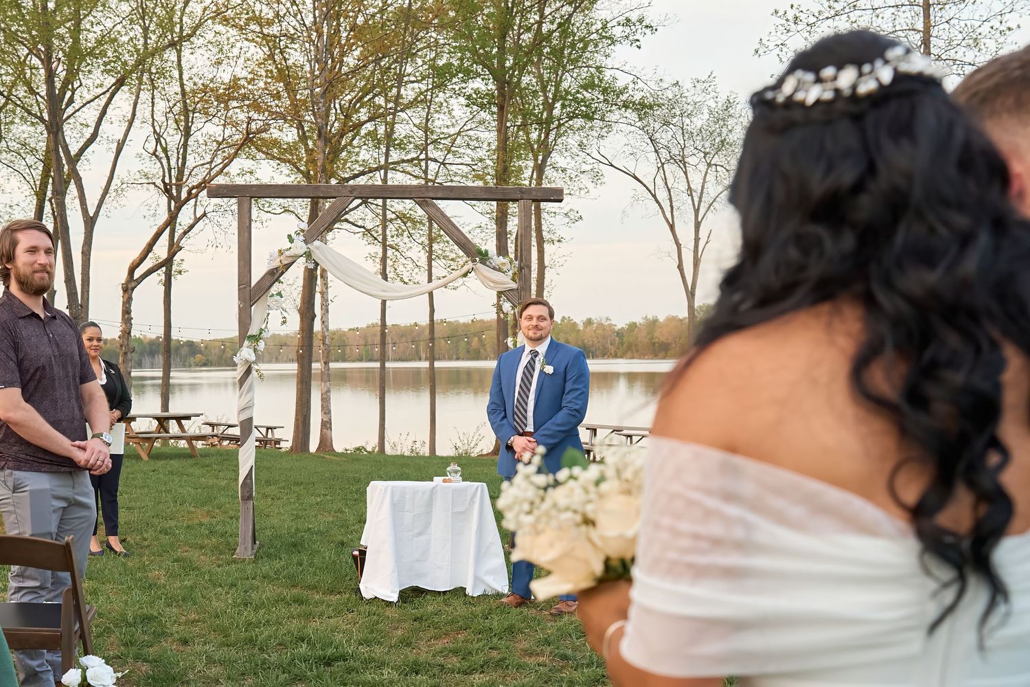 Bride in white off-shoulder gown walks toward groom at outdoor lakeside wedding ceremony with wooden arch backdrop.