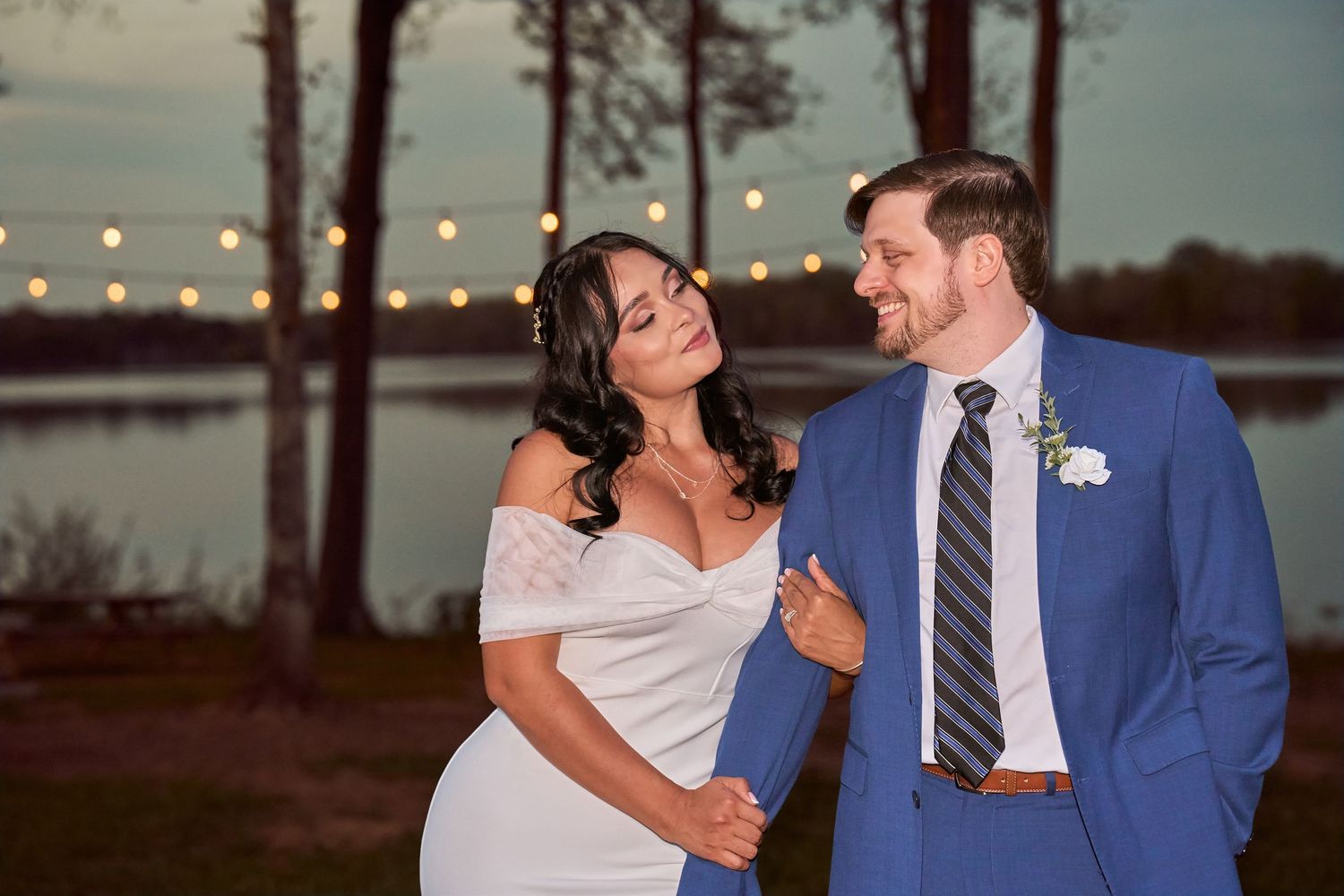 Bride and groom share a romantic glance outdoors at dusk, with string lights and a lake in the background.