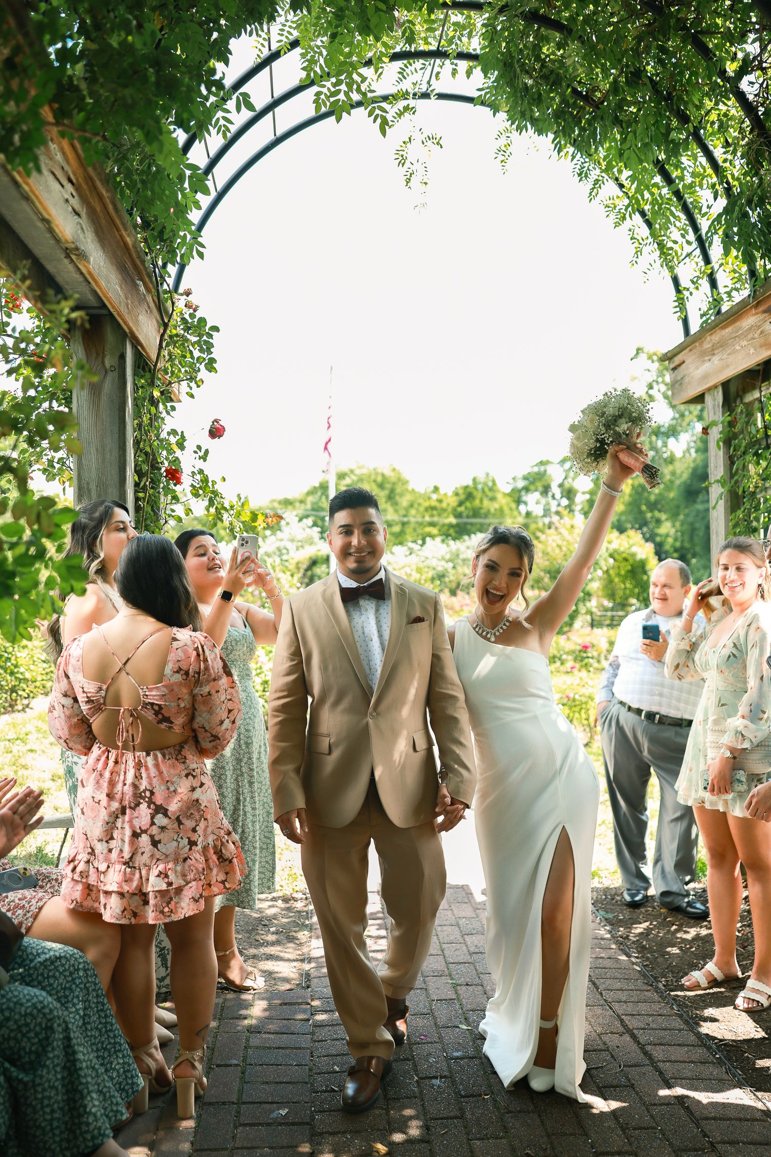 Wedding celebration under garden archway with newlyweds walking down path surrounded by cheering guests.