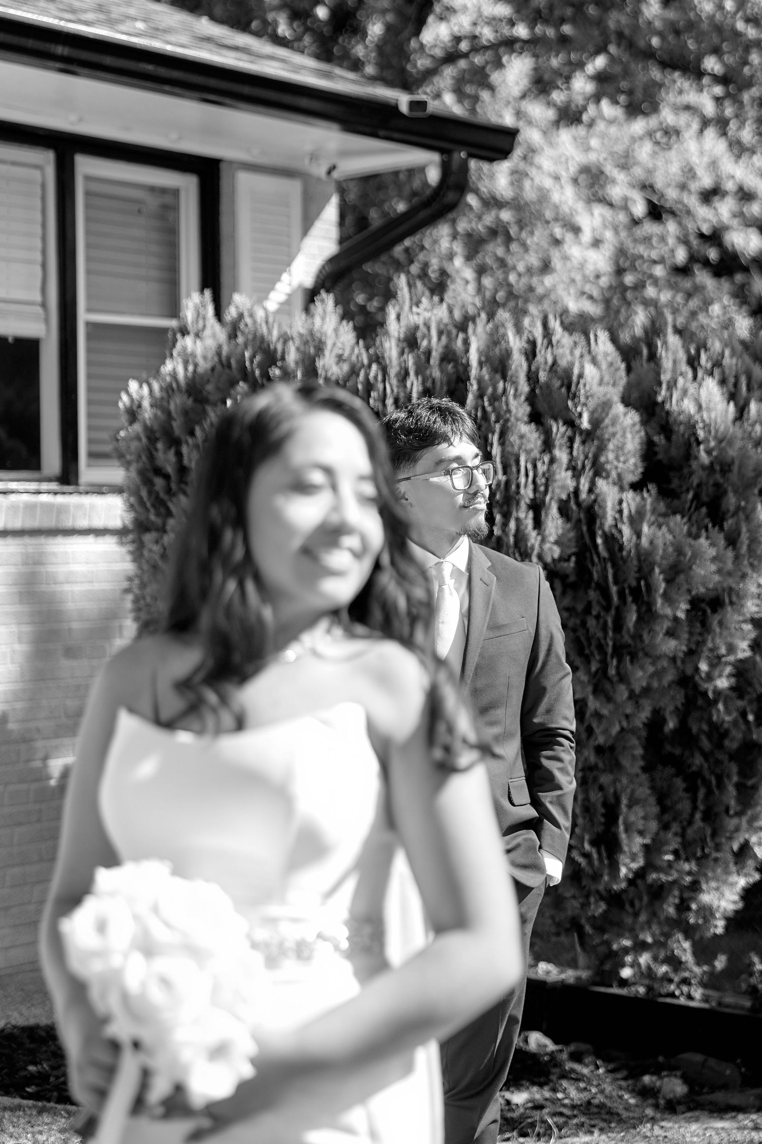 A romantic wedding couple dancing together outdoors in front of a house with greenery.