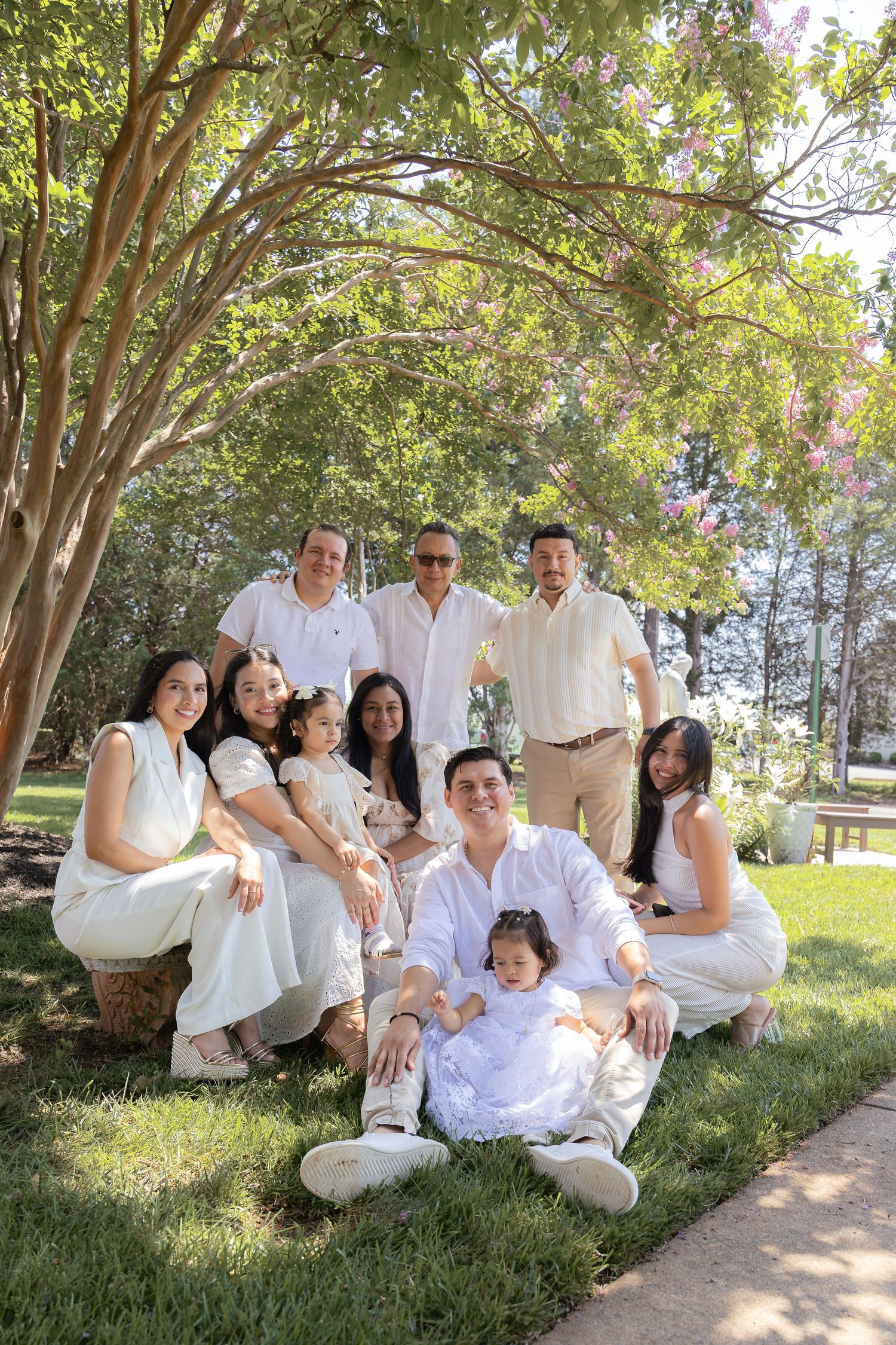 A large family group poses together outdoors in coordinating white outfits under a shady tree.