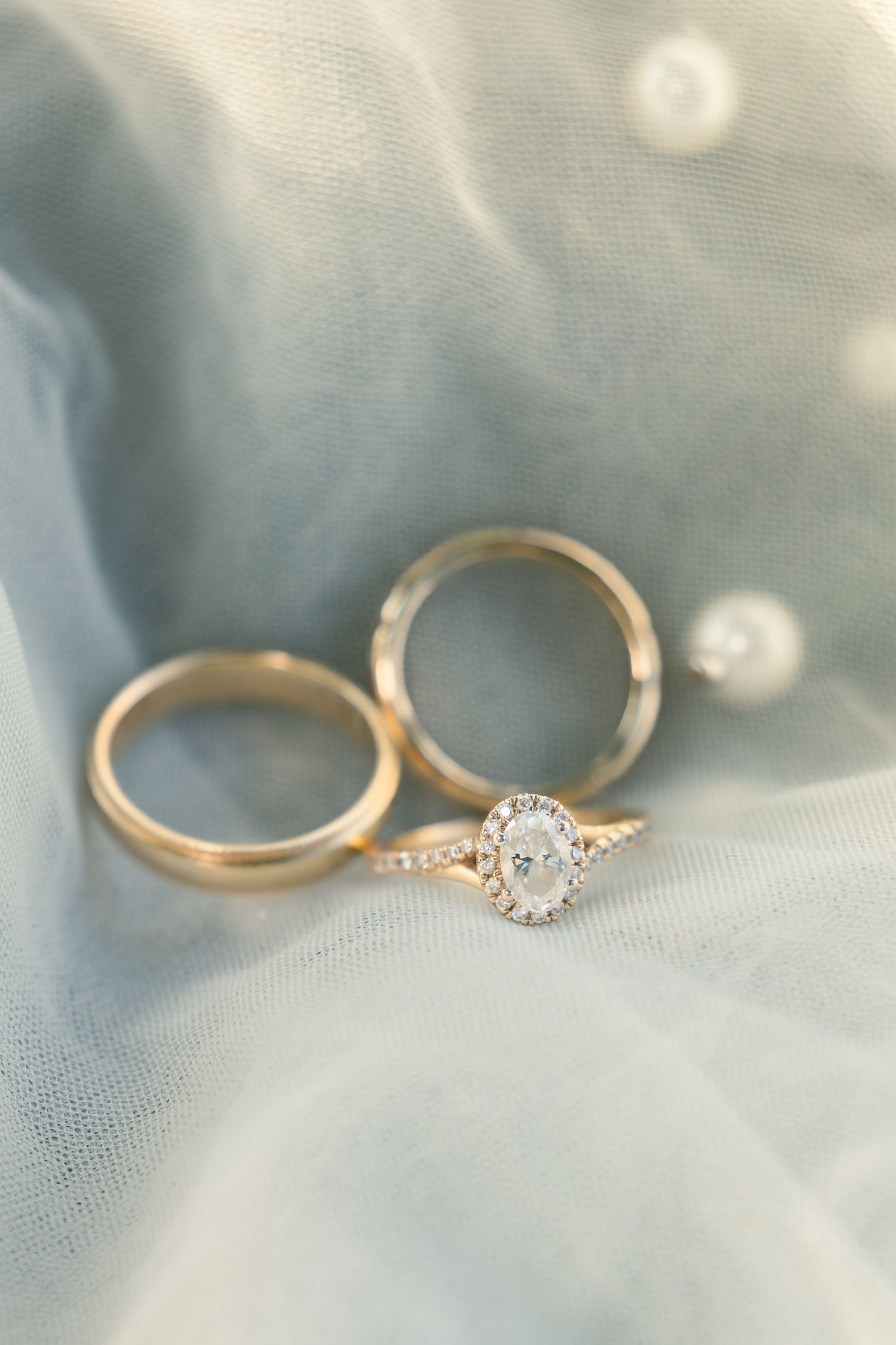 Two elegant gold wedding rings with a sparkling oval diamond engagement ring on light blue fabric background.