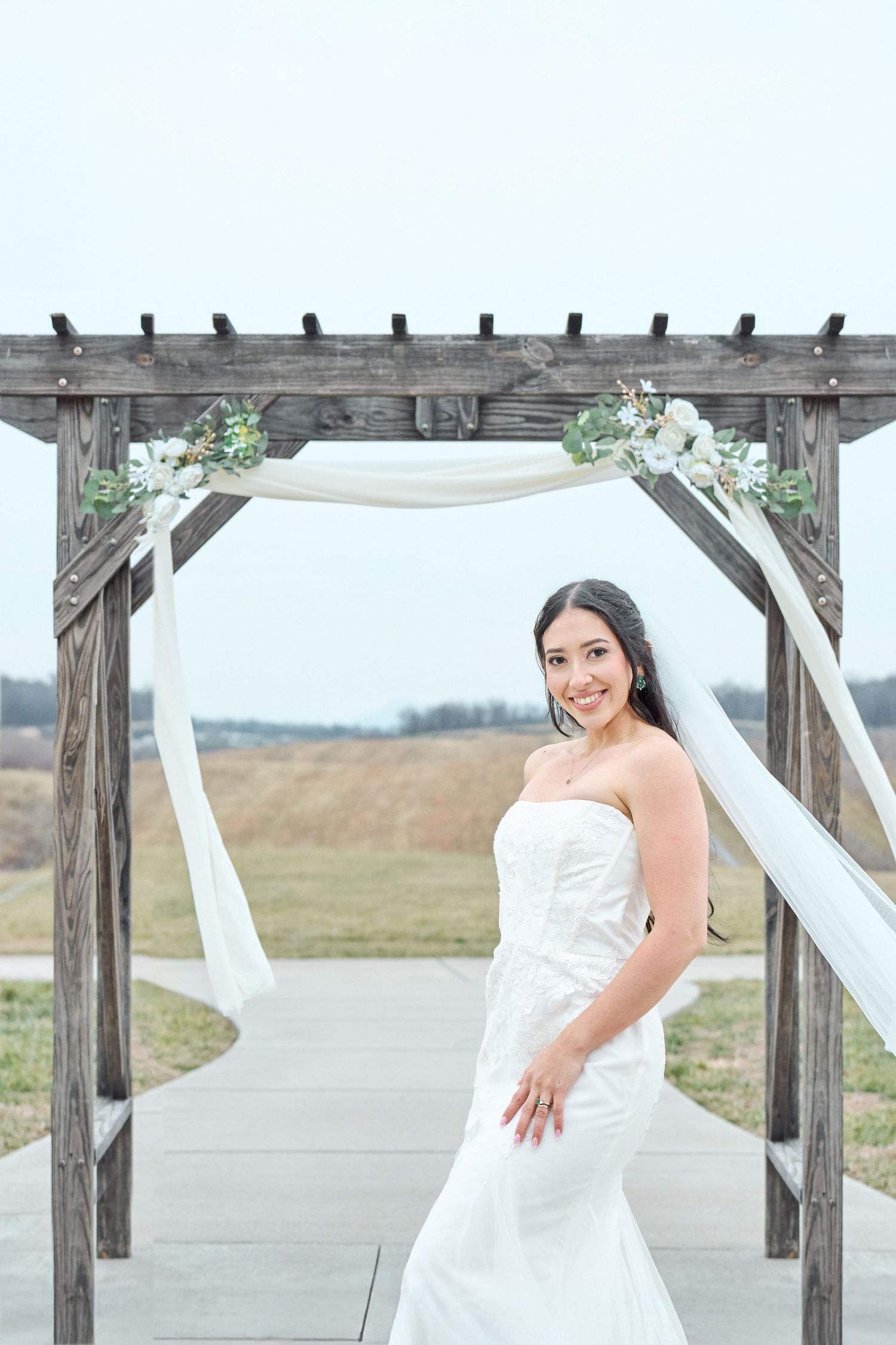Bride in white strapless gown poses under rustic wooden arch decorated with white flowers and fabric.