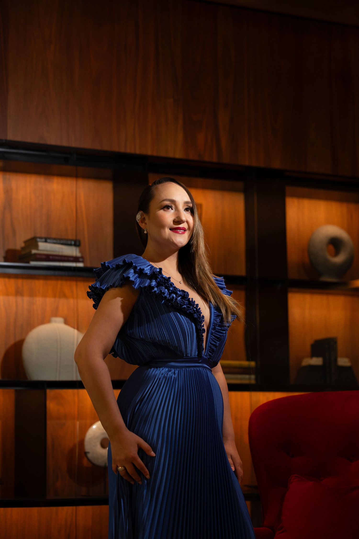 Woman in navy pleated gown poses in warmly lit room with wooden paneled walls.
