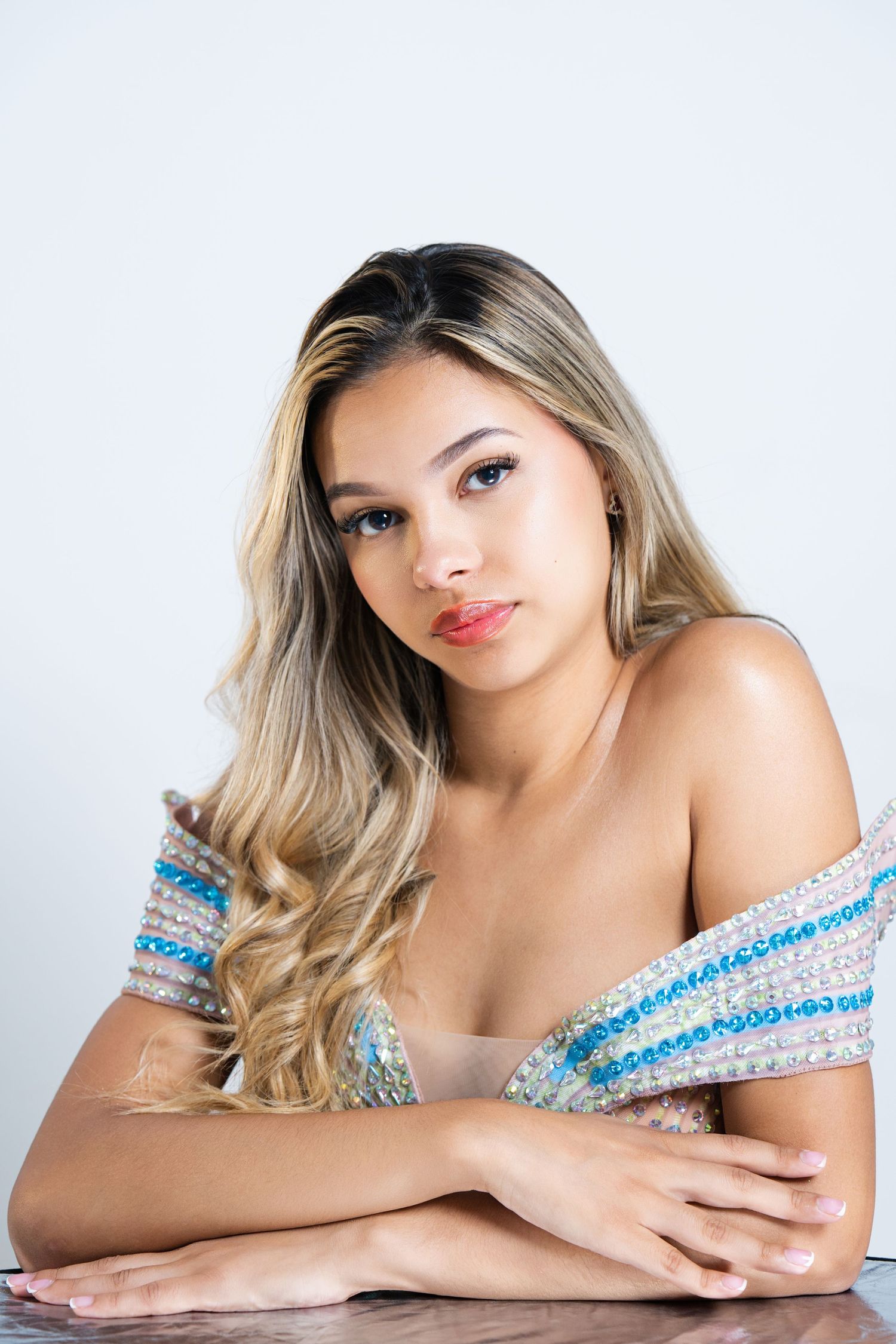 Young woman with long blonde hair wearing a beaded off-shoulder top, posing with arms crossed on a table.