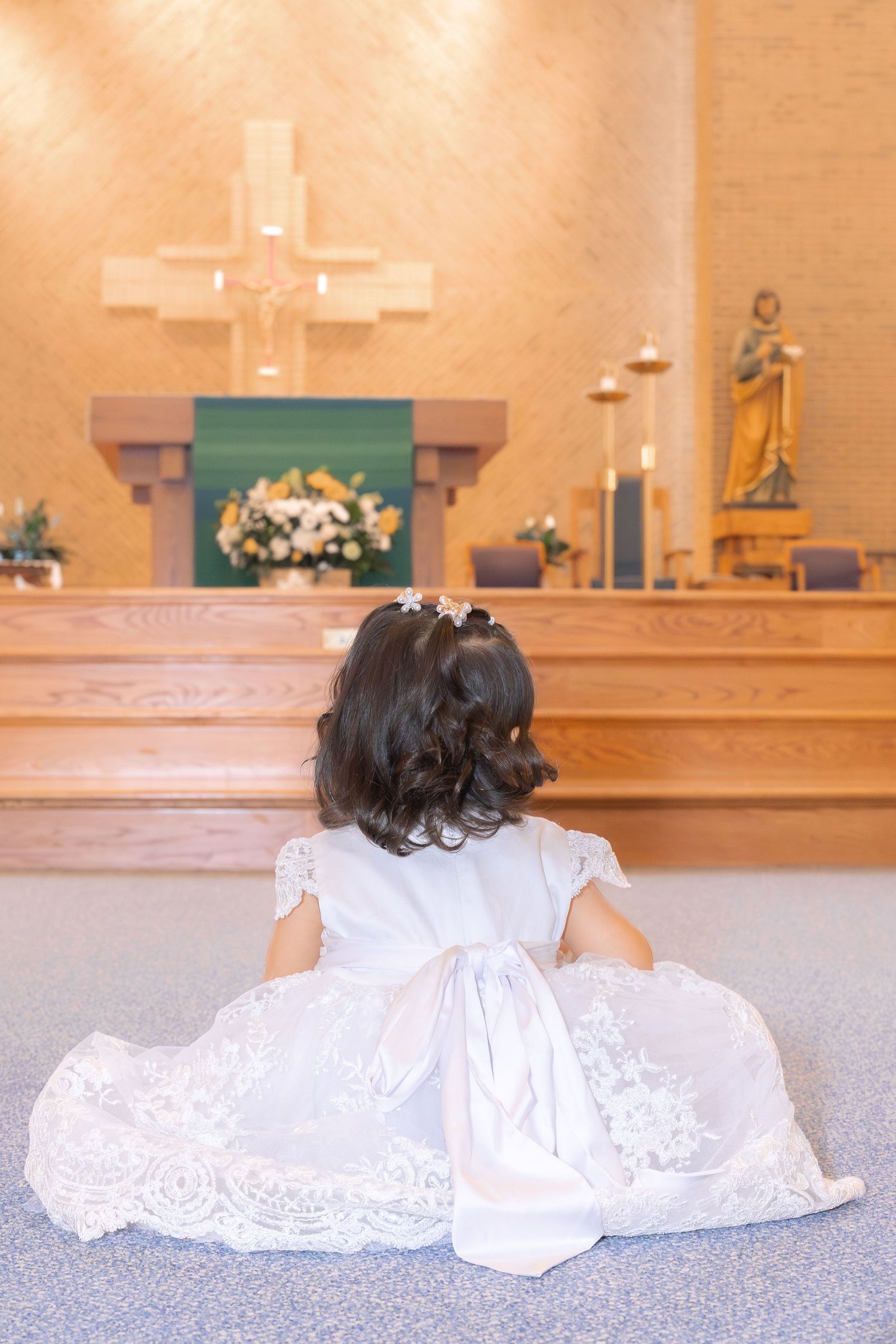 Back view of small child in white lace dress sitting on church floor facing altar.