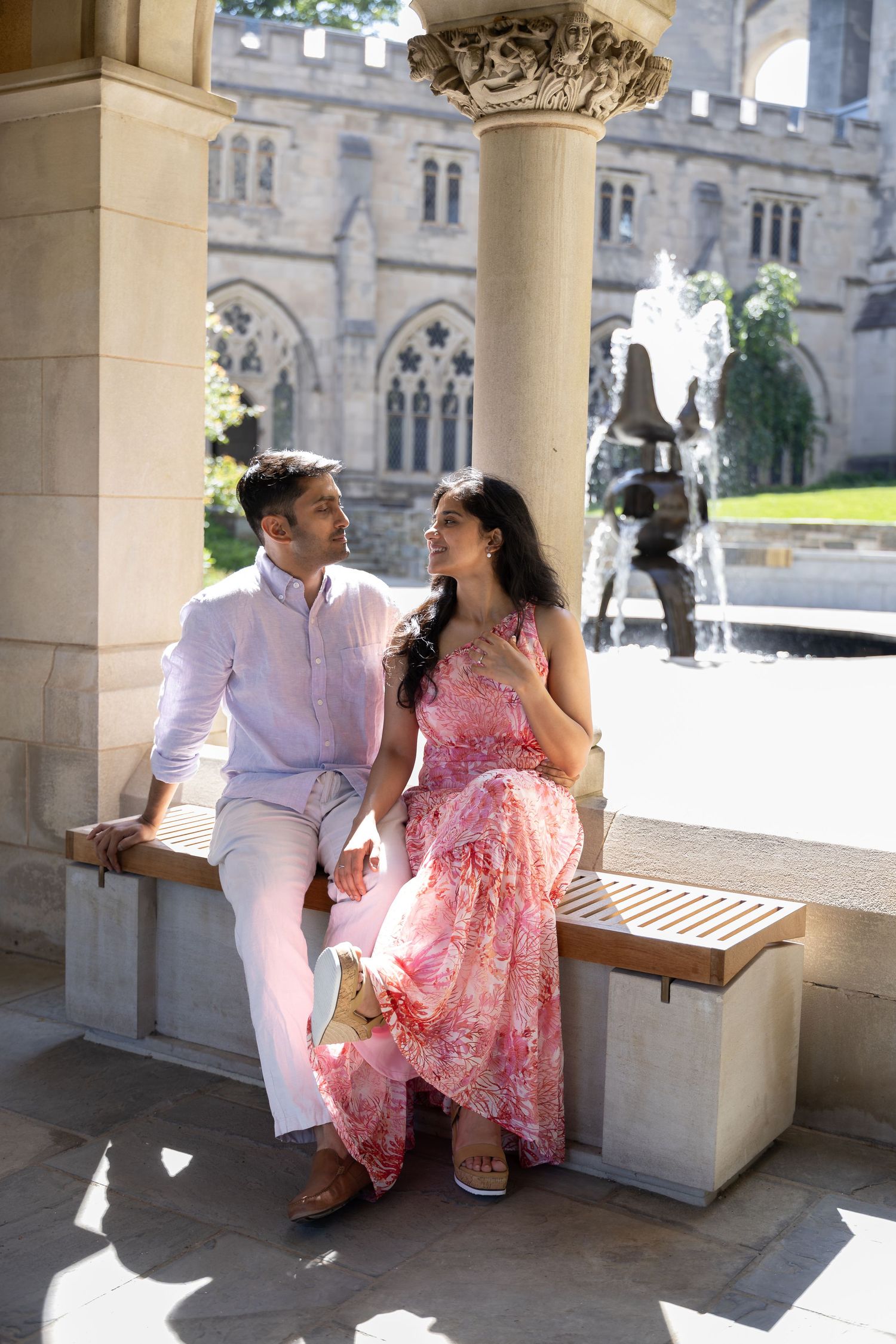 A couple sits on a stone bench in a Gothic architectural setting, with her wearing a pink floral dress and him in light attire.