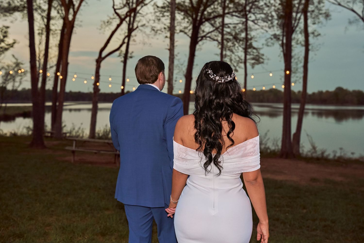 Bride and groom holding hands facing a serene lake at dusk, surrounded by trees with string lights.