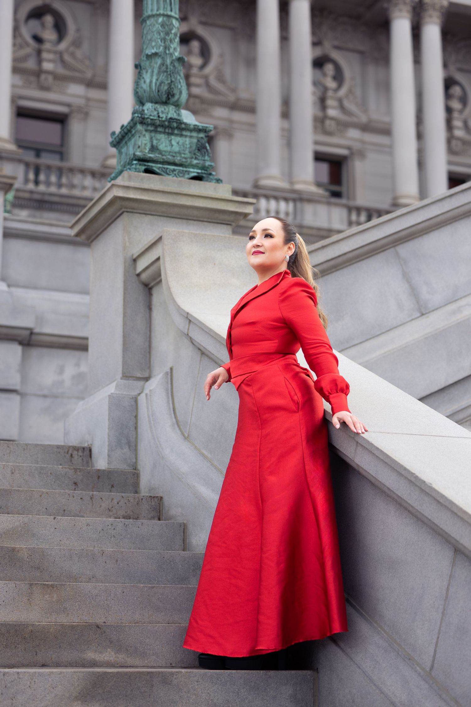 Model in flowing red dress poses elegantly on marble steps of government building.
