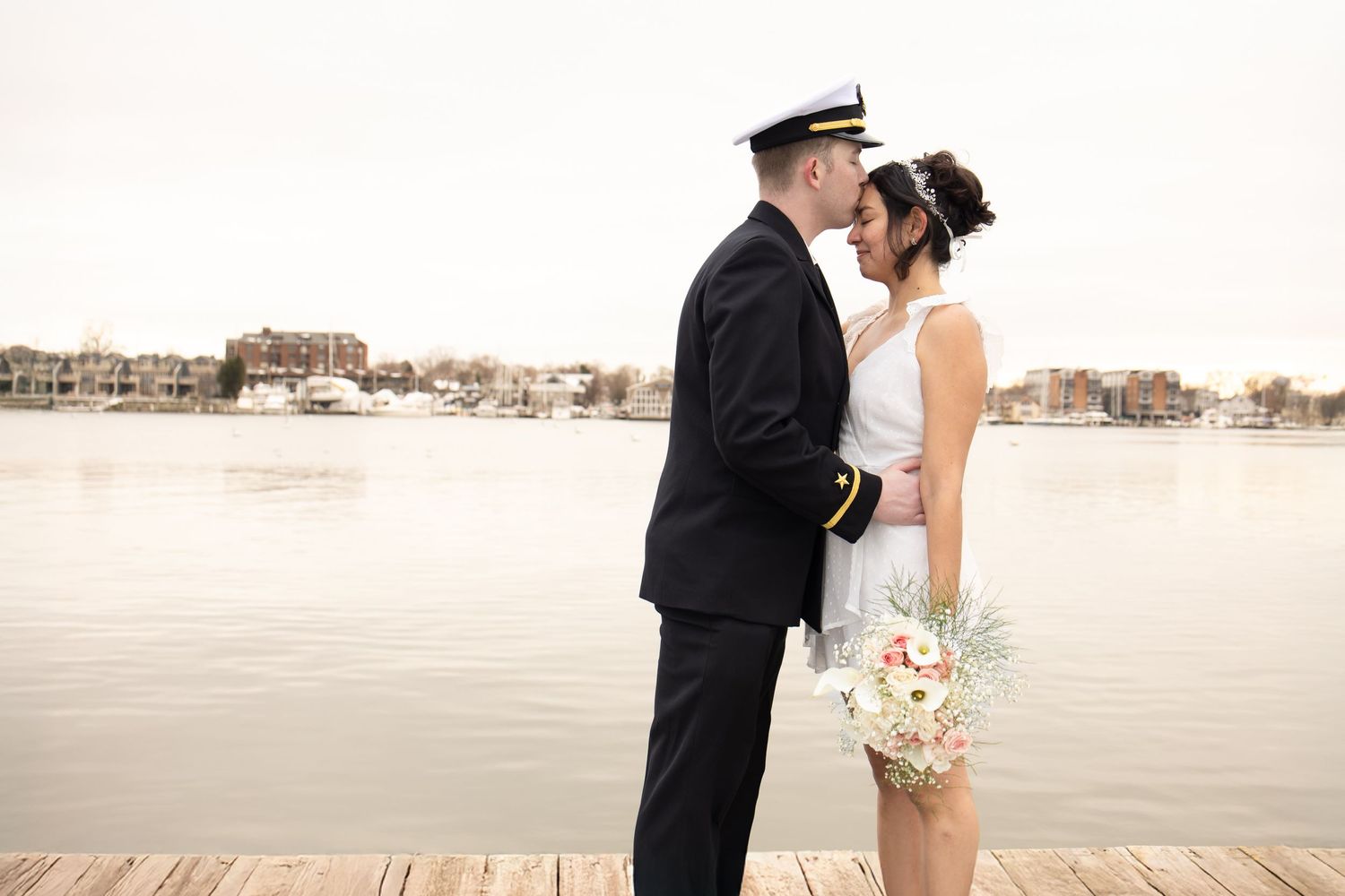 A couple shares a tender moment on a dock with water and buildings in the background.