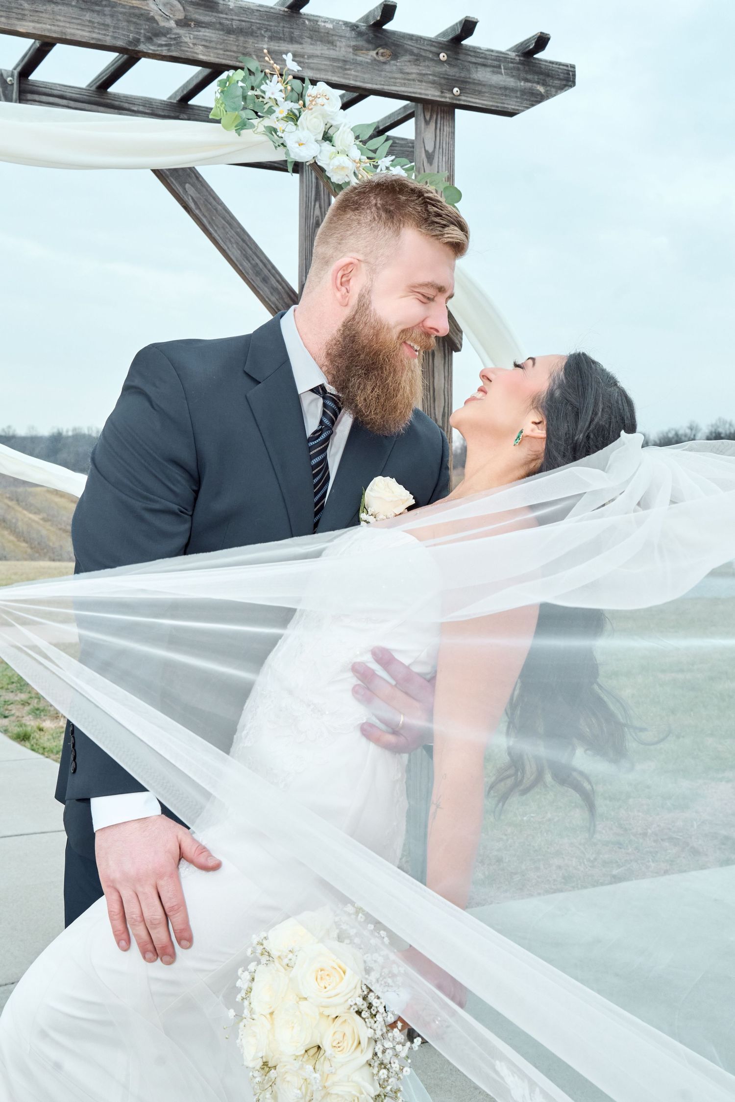 A romantic wedding couple embraces under a wooden pergola with flowing white veil and floral decorations.