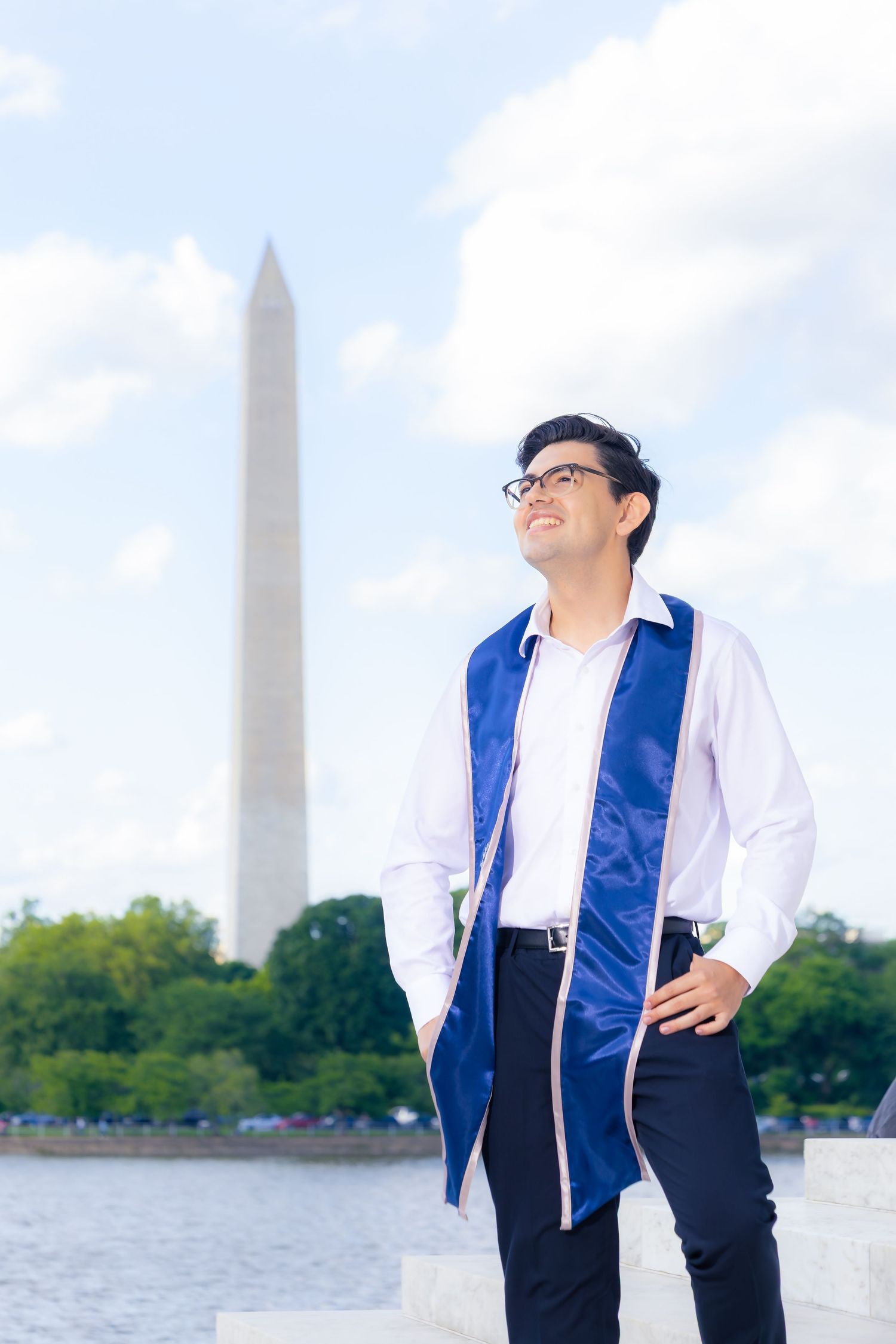 Graduate in blue and white regalia poses next to Washington Monument at Jefferson Memorial.