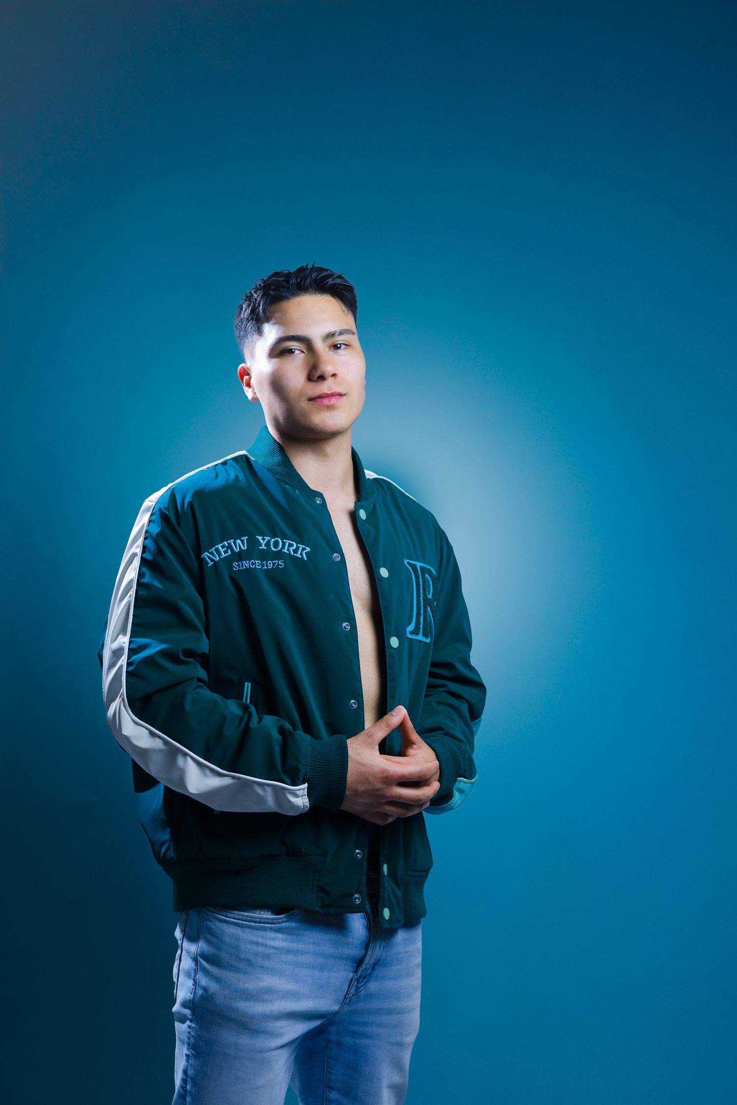 Someone poses confidently in a navy blue varsity jacket and jeans against a vibrant blue studio backdrop with dramatic lighting.