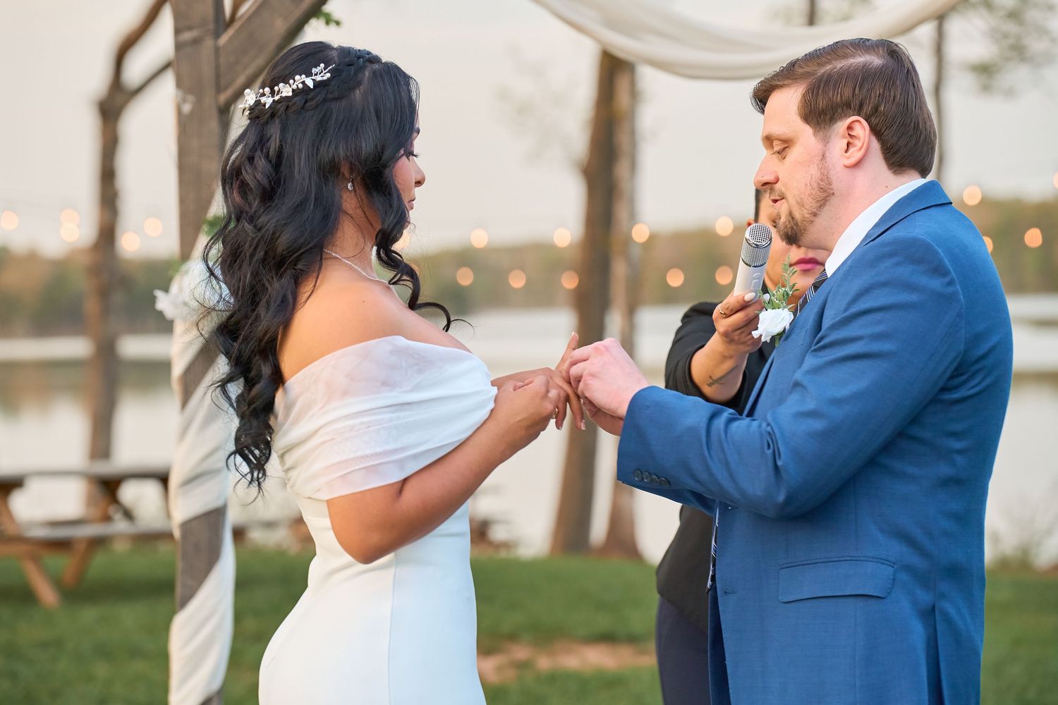 Bride placing wedding ring on groom's finger during outdoor ceremony near lake at sunset.