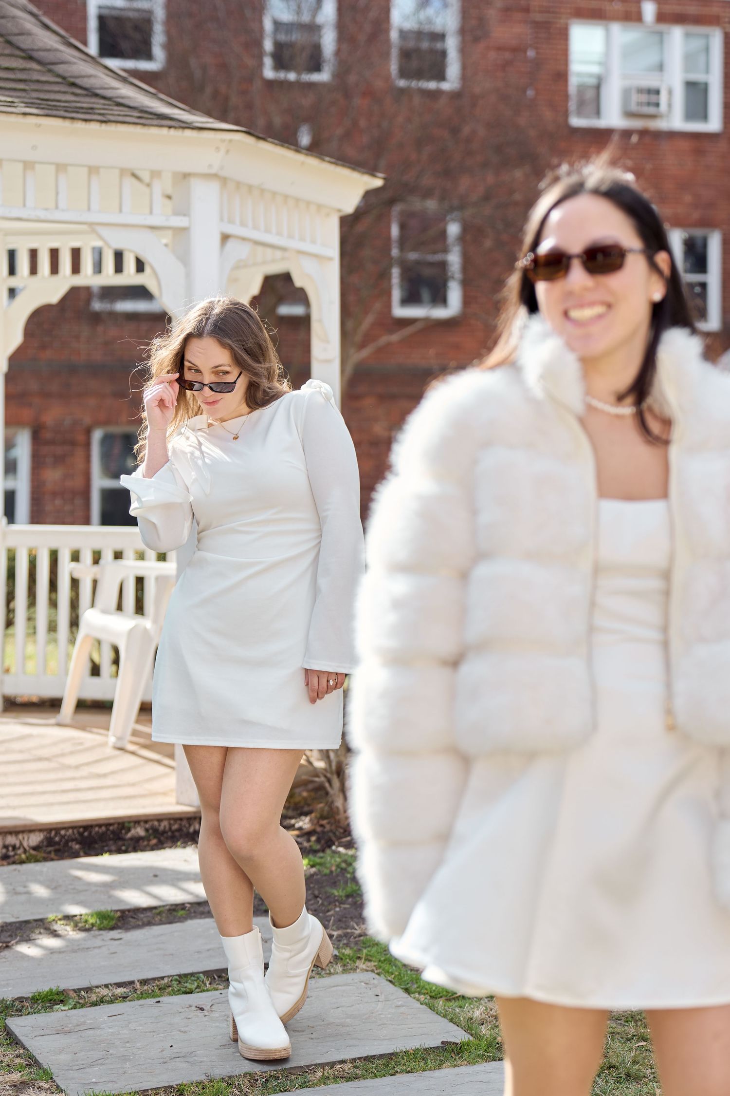 Women in coordinated white outfits pose along stone pathway near brick building and white gazebo.