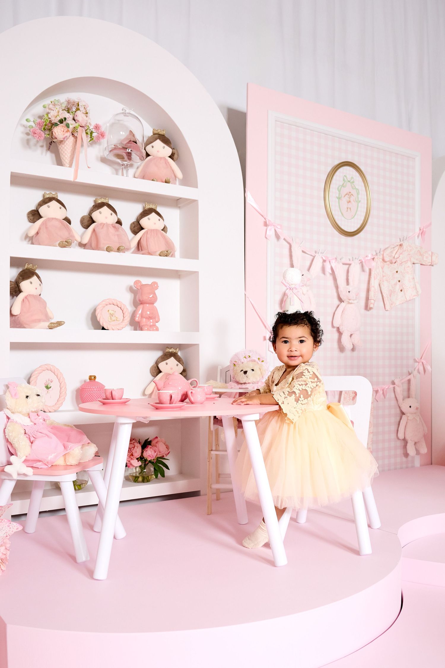 Toddler girl in yellow tutu dress stands at pink table with dolls and toys in a pink themed party room.