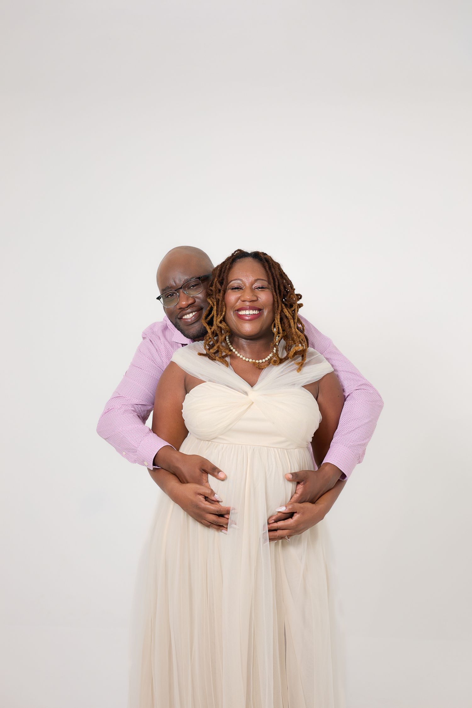 Happy couple posing for maternity photo, man embracing pregnant woman in cream gown, white studio background.