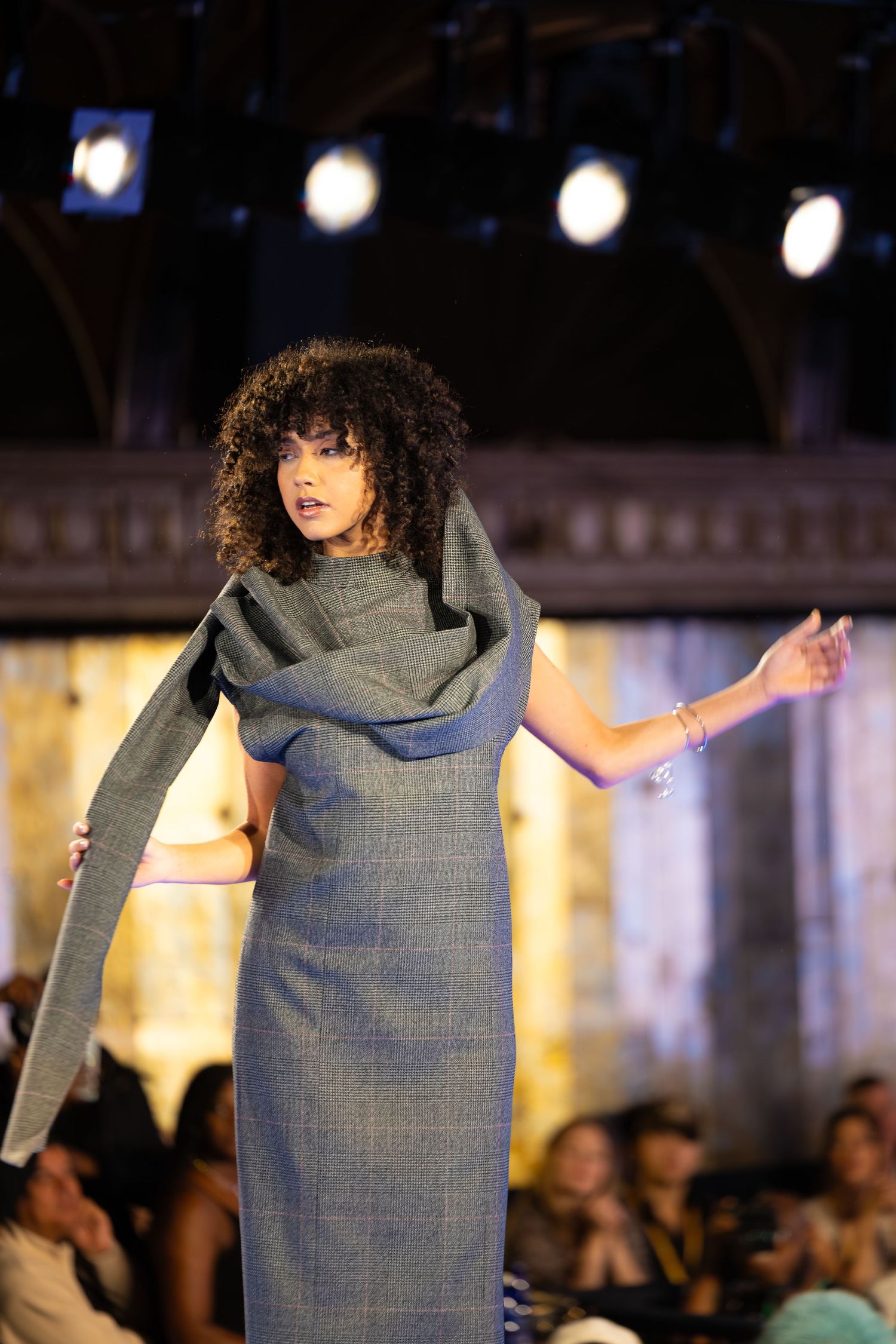 Model in grey structured dress with dramatic draped detail performs on fashion runway.