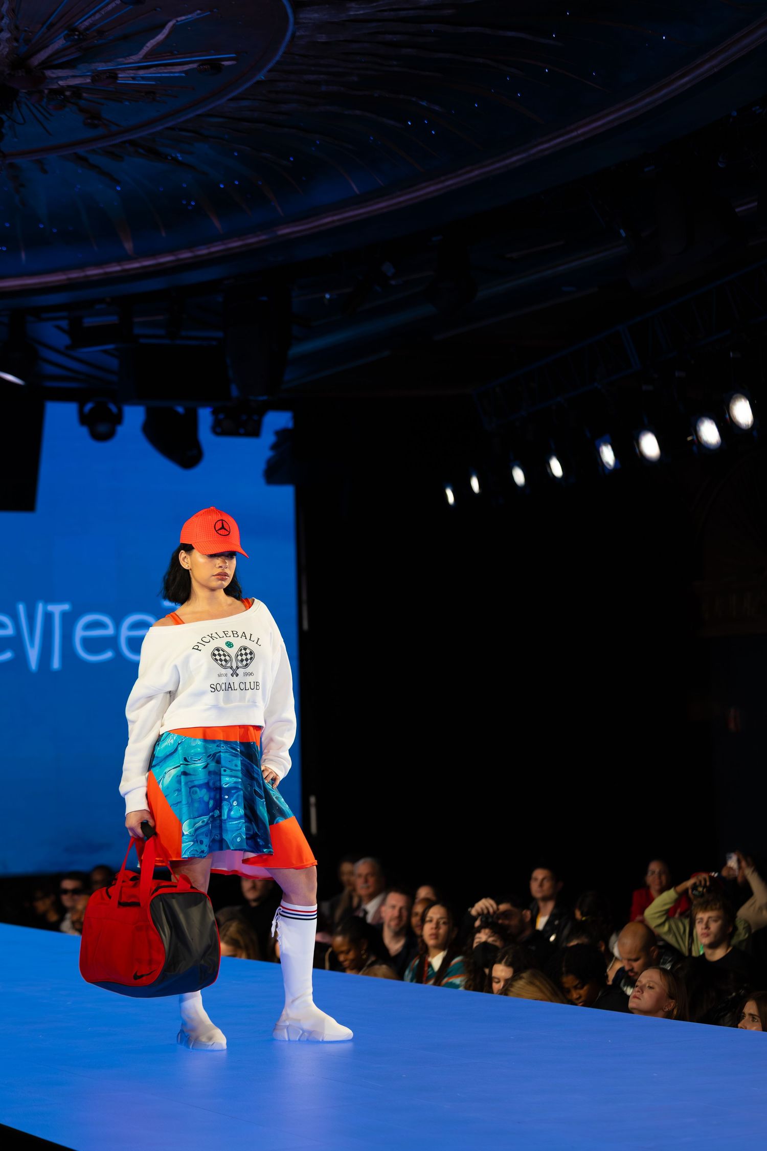 Casual streetwear look with white graphic sweater and red accessories on blue-lit runway.