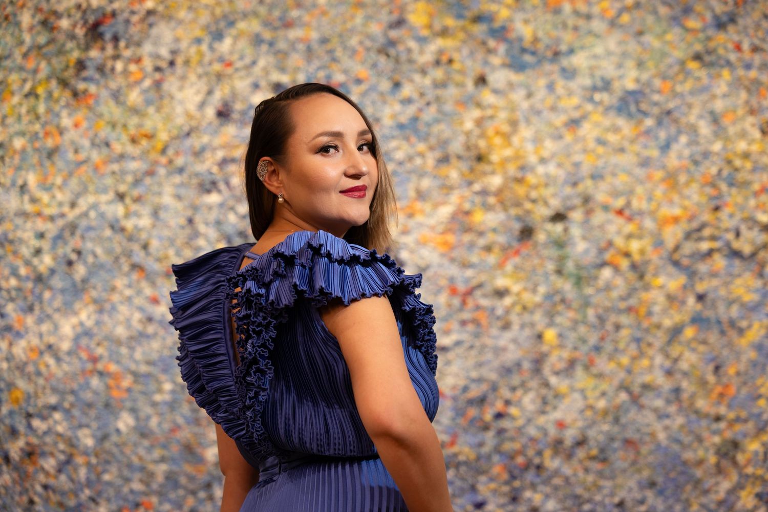 Woman in elegant navy pleated dress poses against colorful abstract wall background during fashion photoshoot.