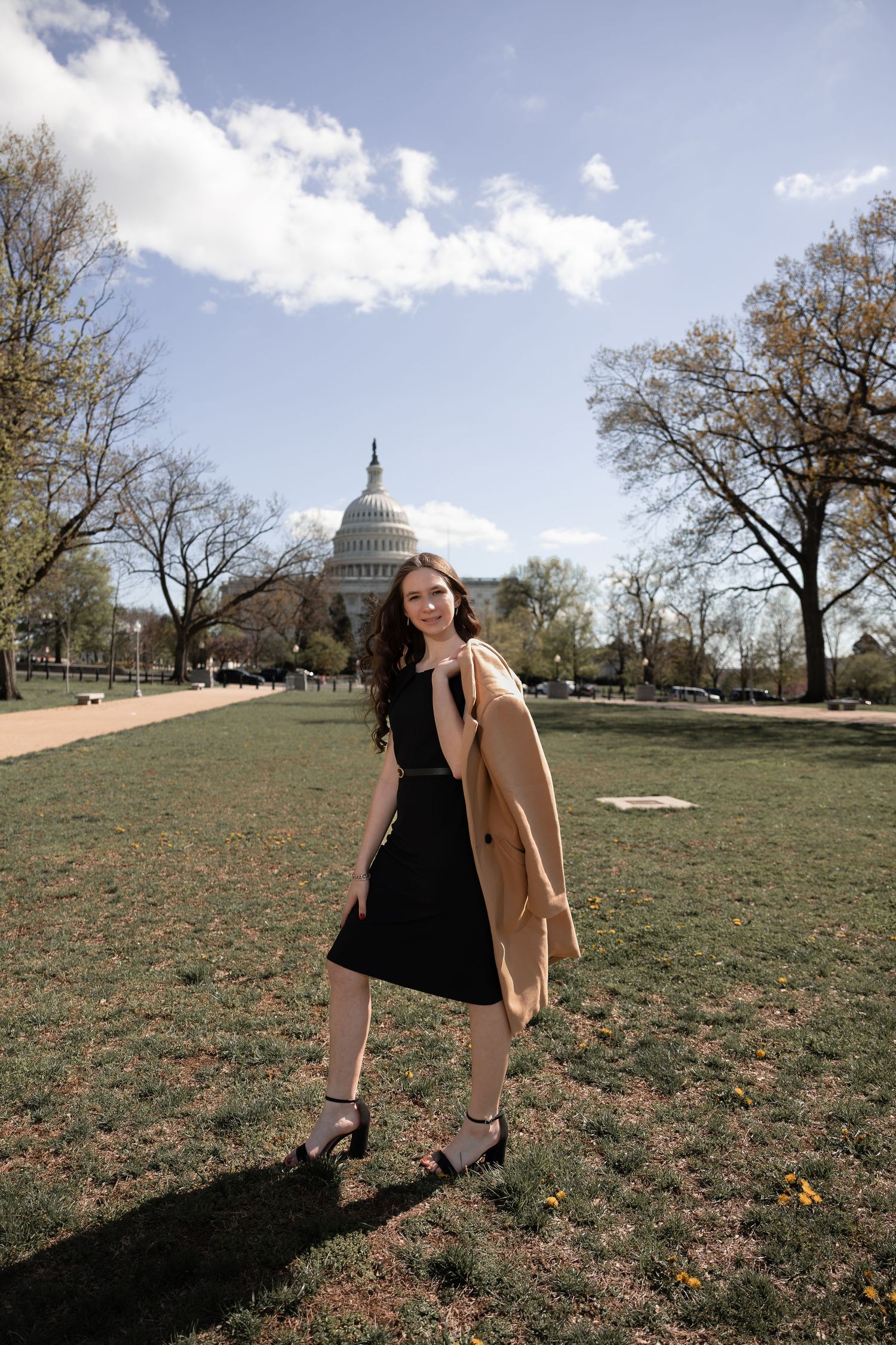 A person in a black dress and tan coat poses on the National Mall with the US Capitol building visible in the background.