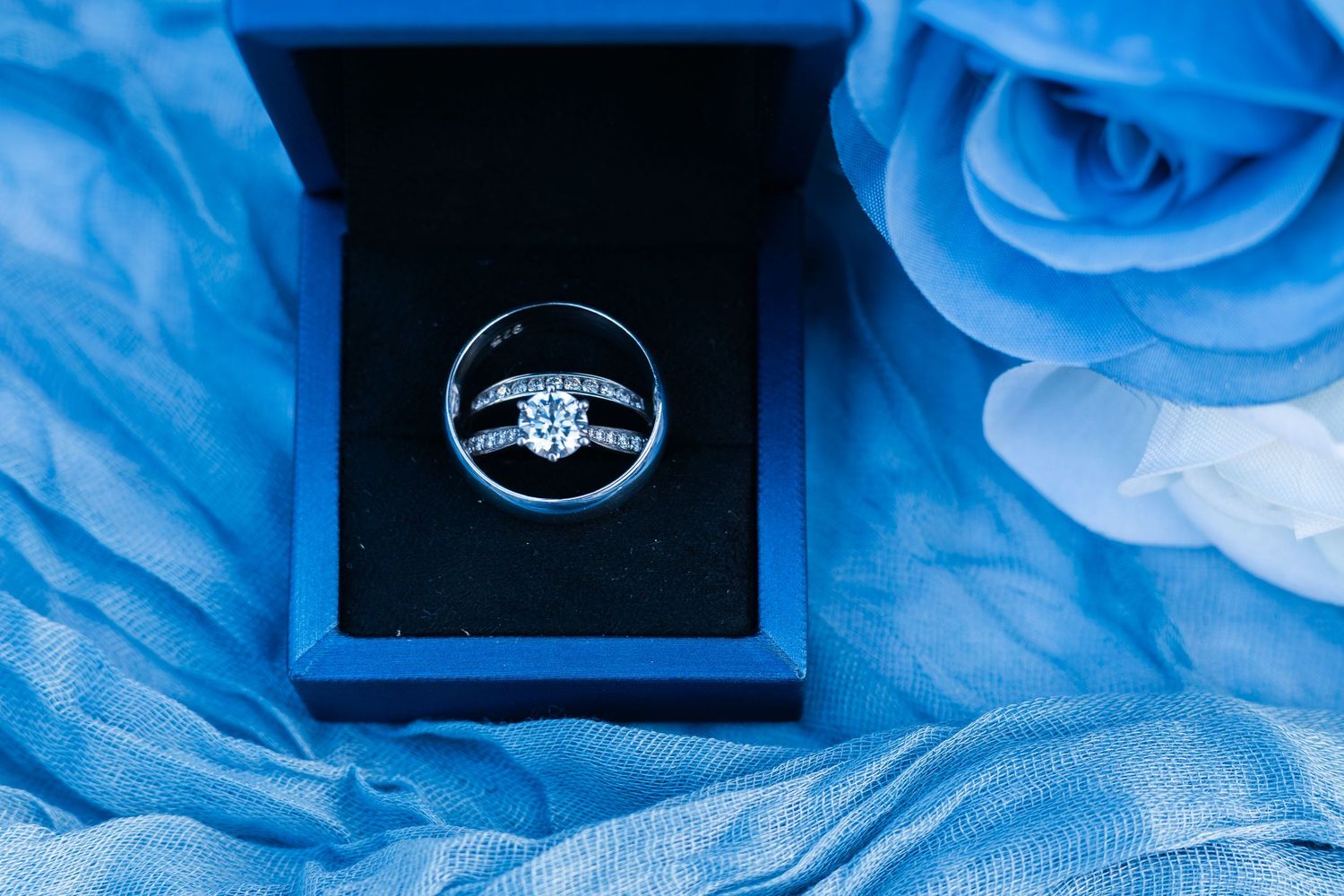 Diamond engagement ring in navy blue box surrounded by blue tulle fabric and roses.