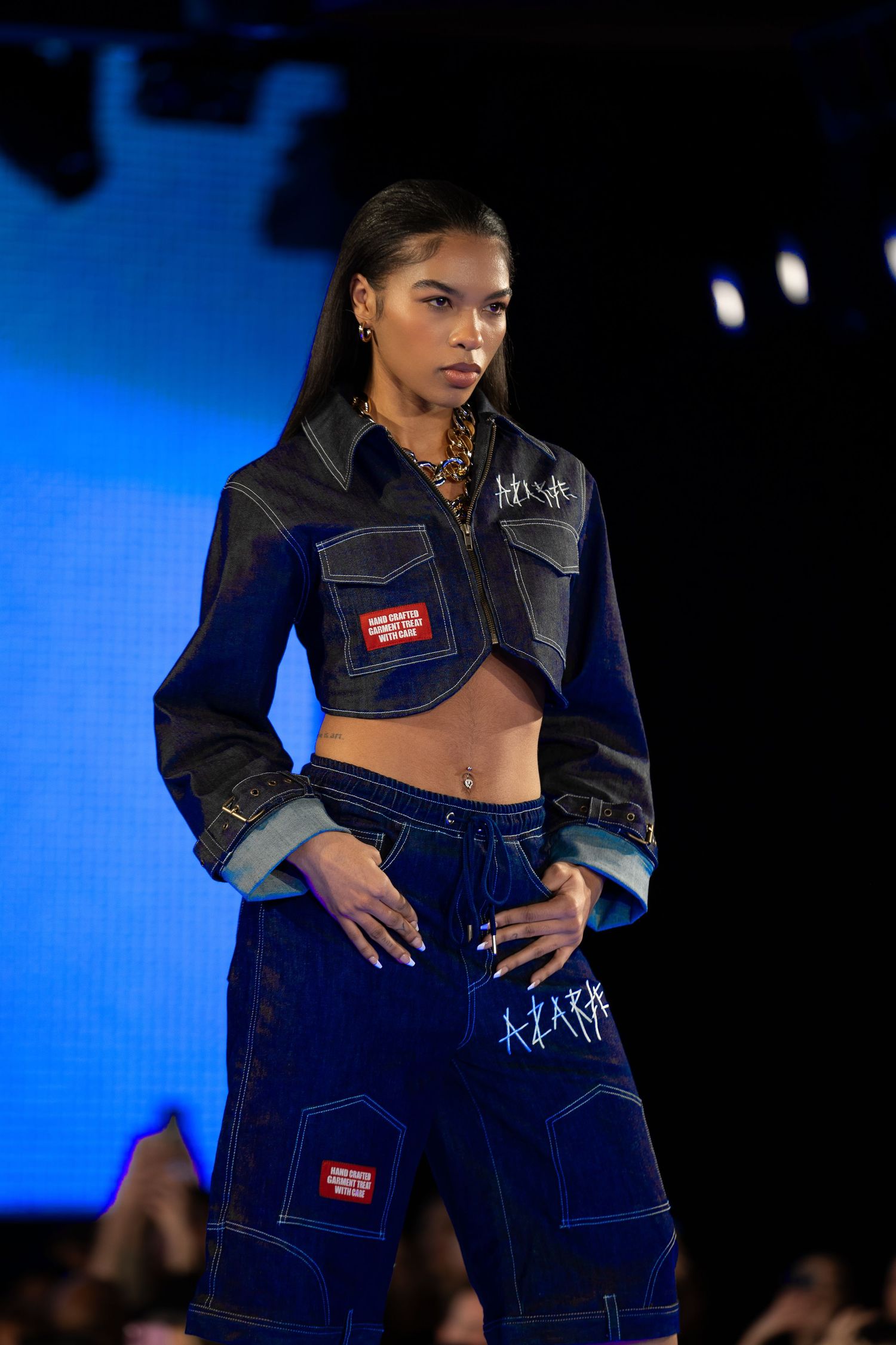 Model showcases cropped denim jacket and matching pants with patches on blue-lit catwalk.