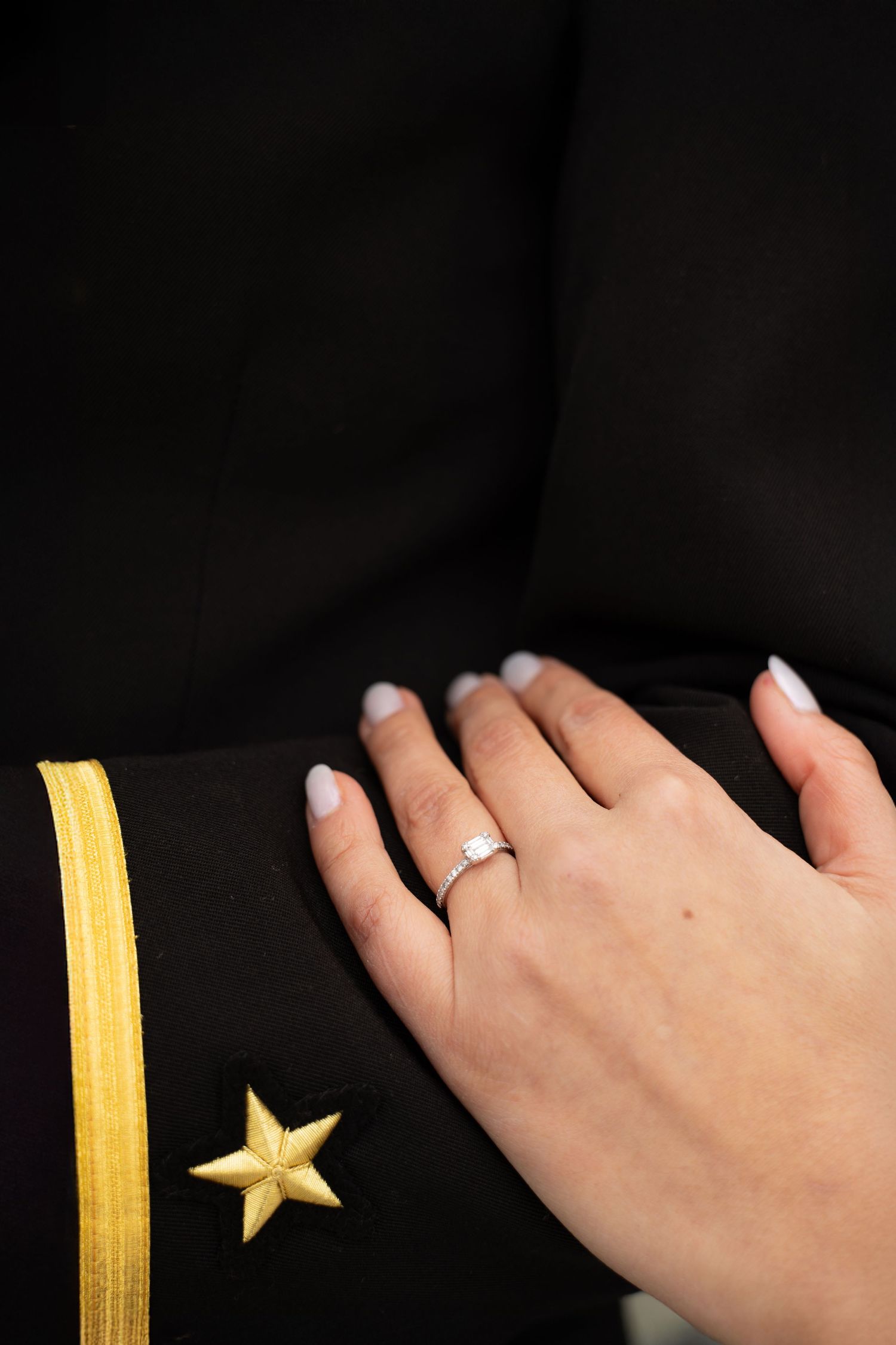 A close-up of an engagement ring on a hand against a naval uniform with gold stripes.