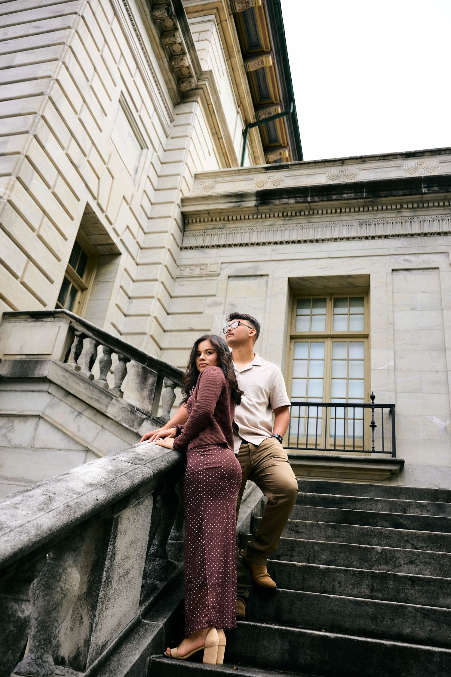 Romantic couple poses on grand stone steps of historic building in vintage-inspired fashion photoshoot.