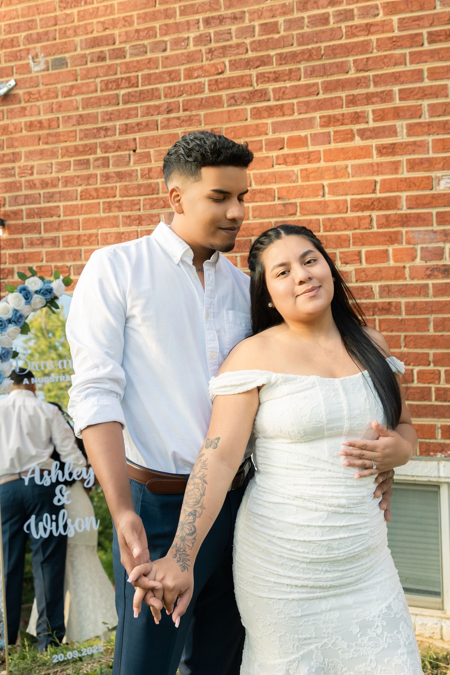 A couple in wedding attire embraces against a brick wall backdrop during an outdoor photoshoot.