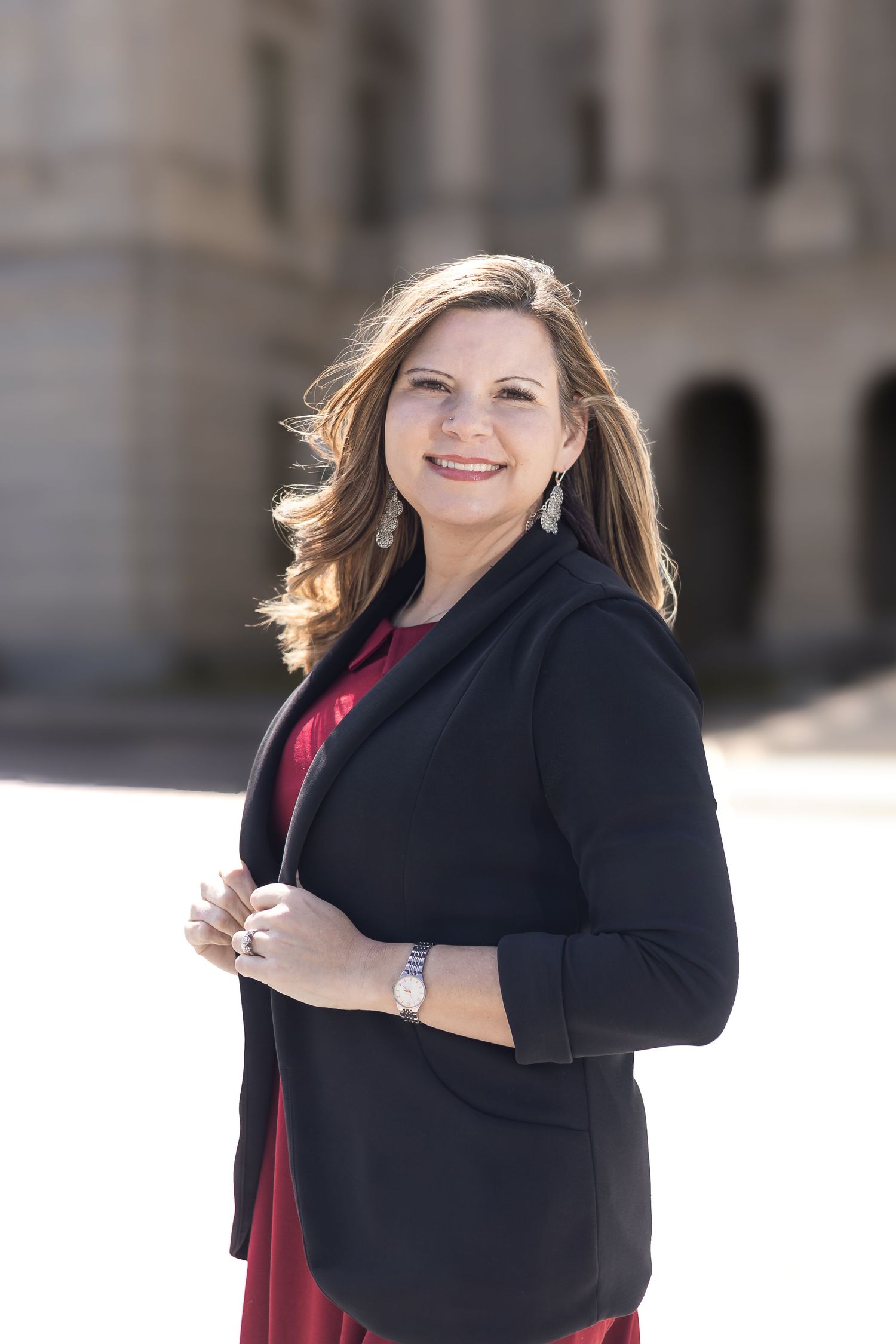Professional headshot of a business person wearing a black blazer and red dress against a blurred architectural background.