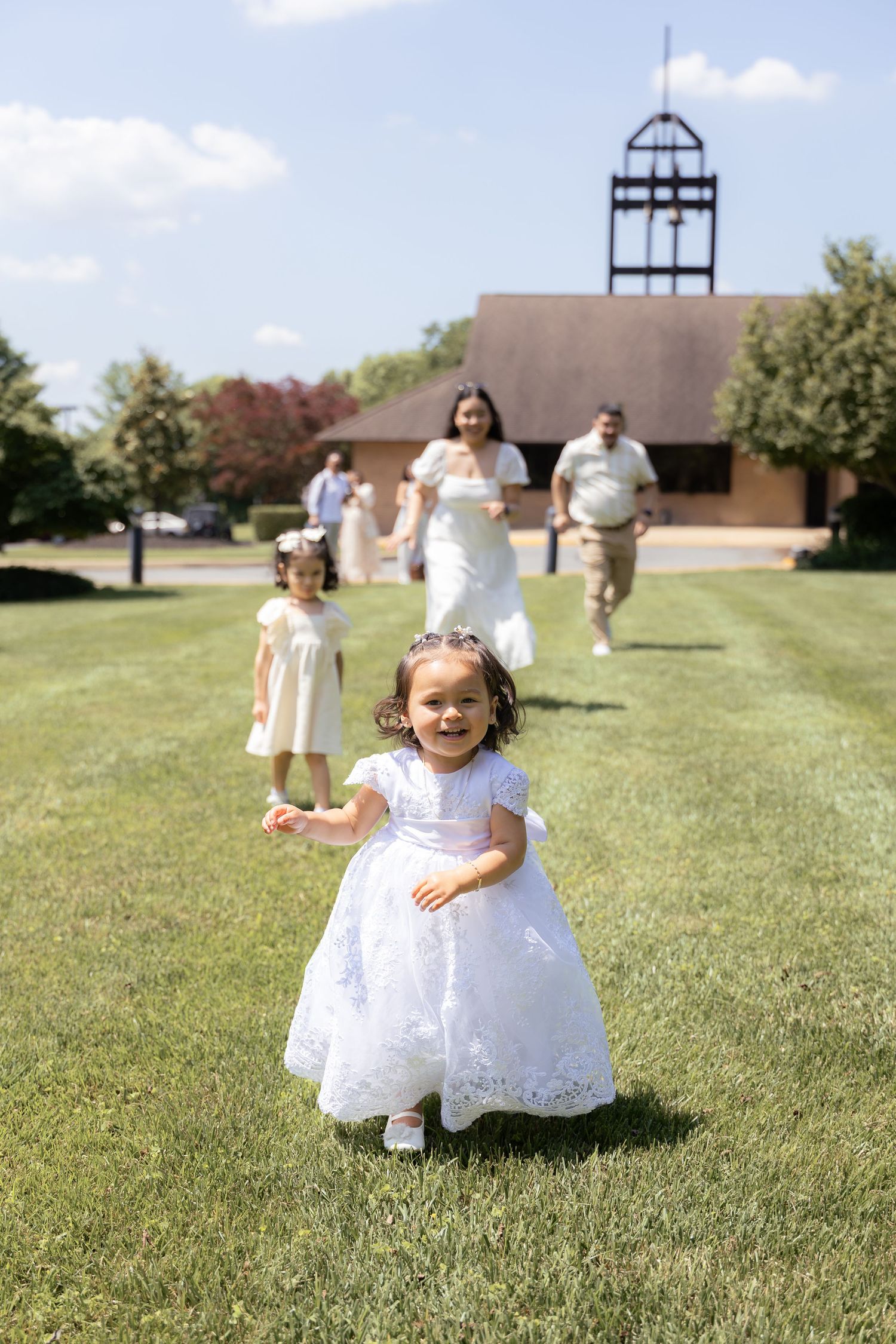 A group of children in white dresses playing and running on a grassy field with a church building in the background.