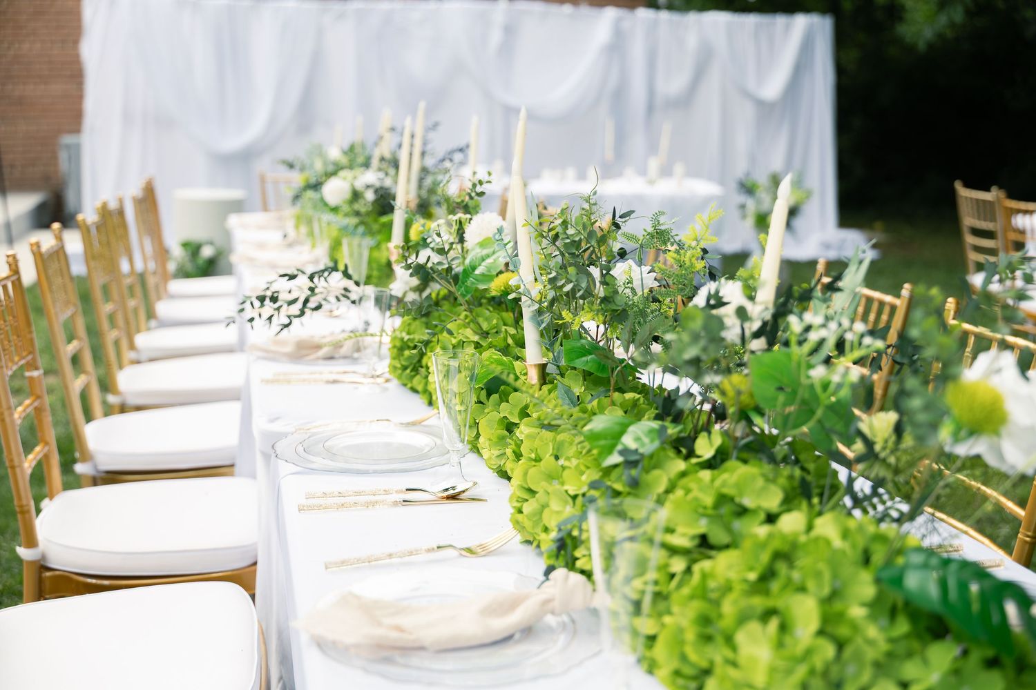 Elegant wedding reception table setting with white candles and lush green foliage runner down long white tablecloth.