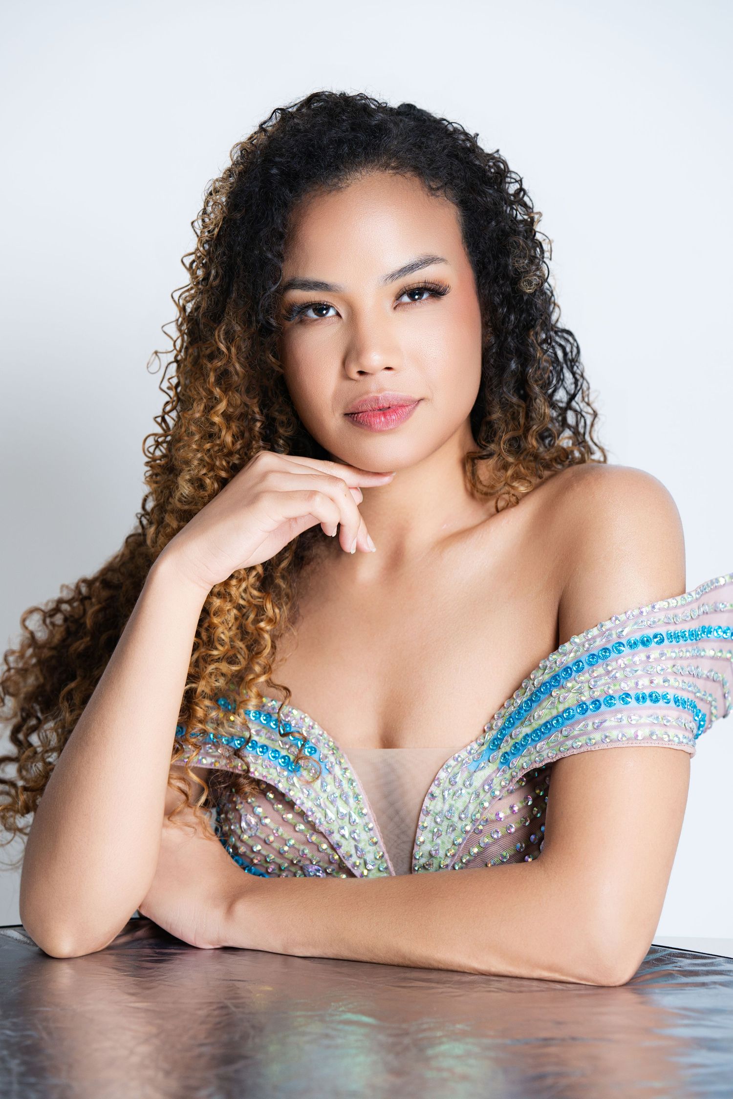 Young woman with curly hair in a sparkling off-shoulder gown poses elegantly at a table against a white background.