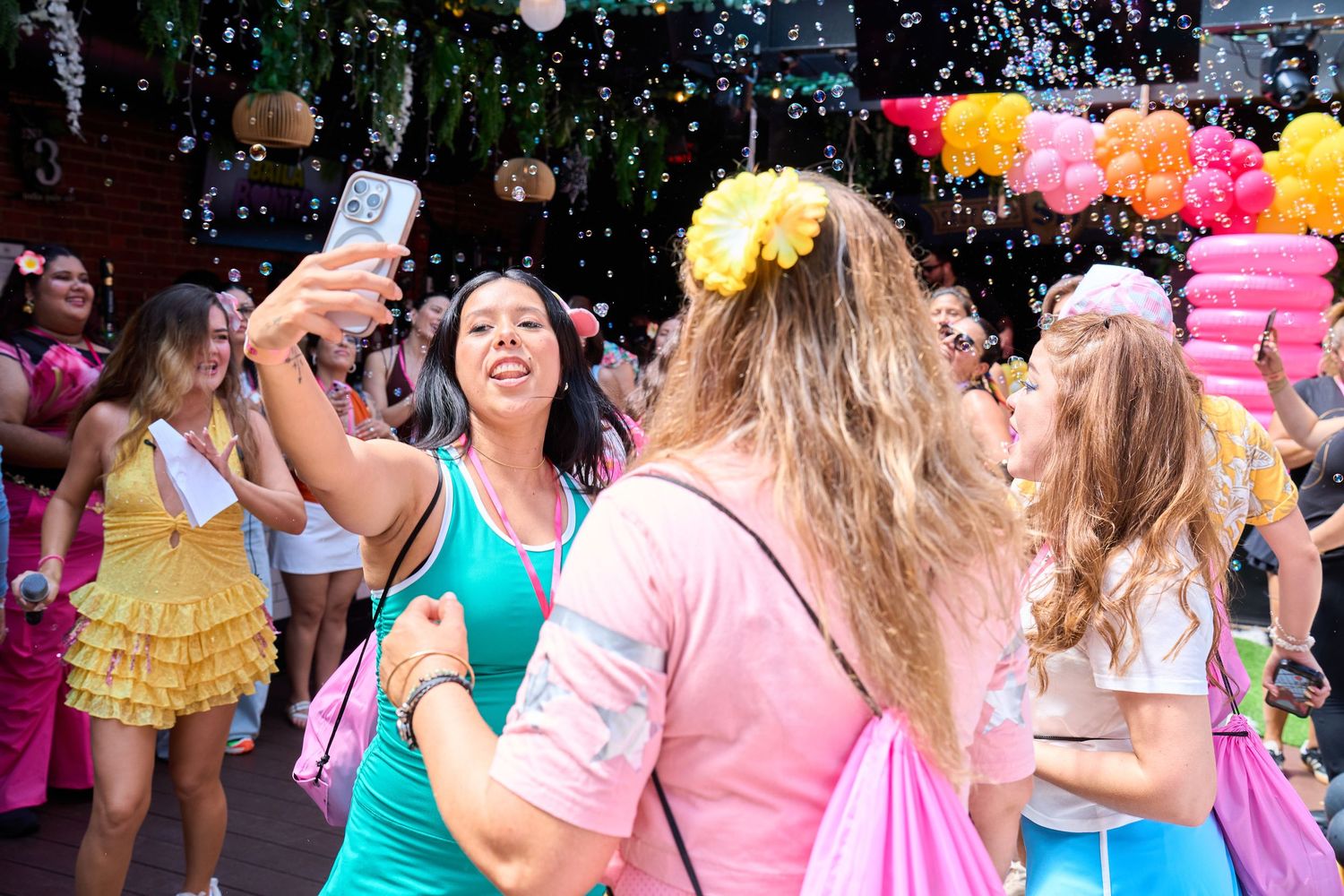 Women in colorful outfits celebrate outdoors with bubbles, one taking a selfie at a festive party event.