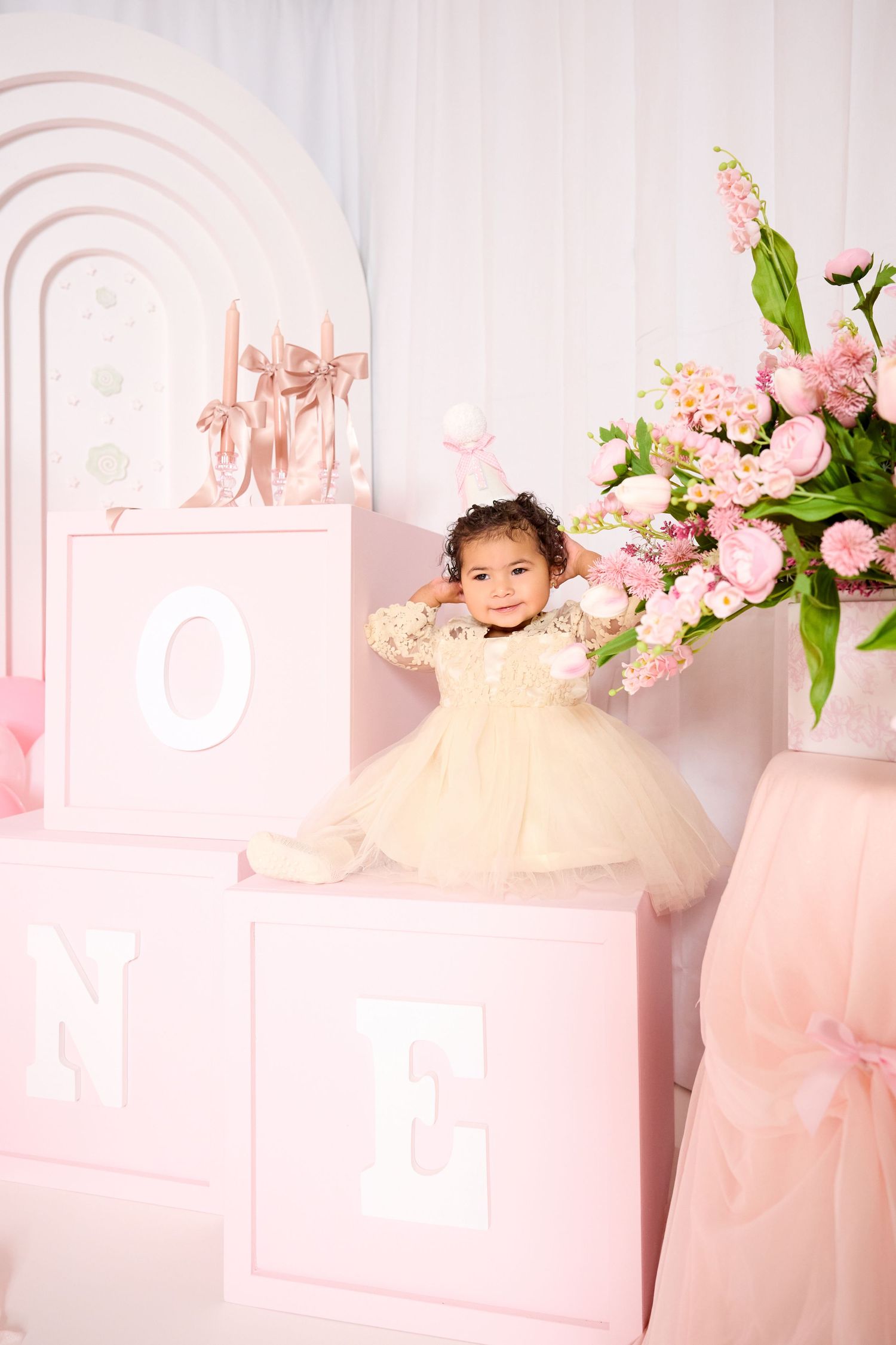Toddler girl in cream tutu dress poses with pink ONE birthday blocks and floral arrangements at first birthday party.