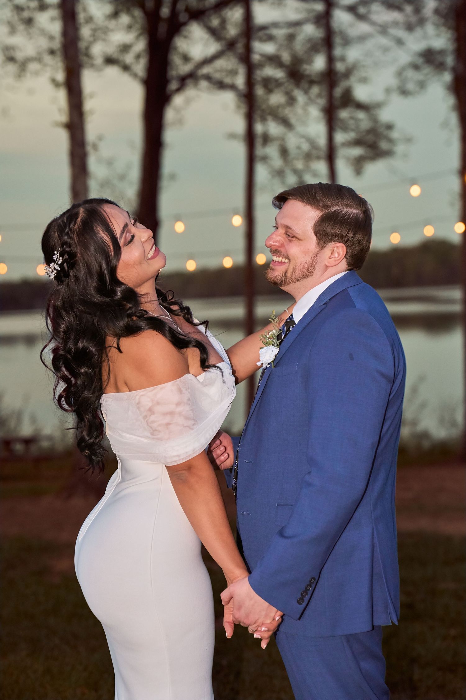 Bride in white off-shoulder gown and groom in blue suit hold hands smiling at sunset by a lake with string lights.