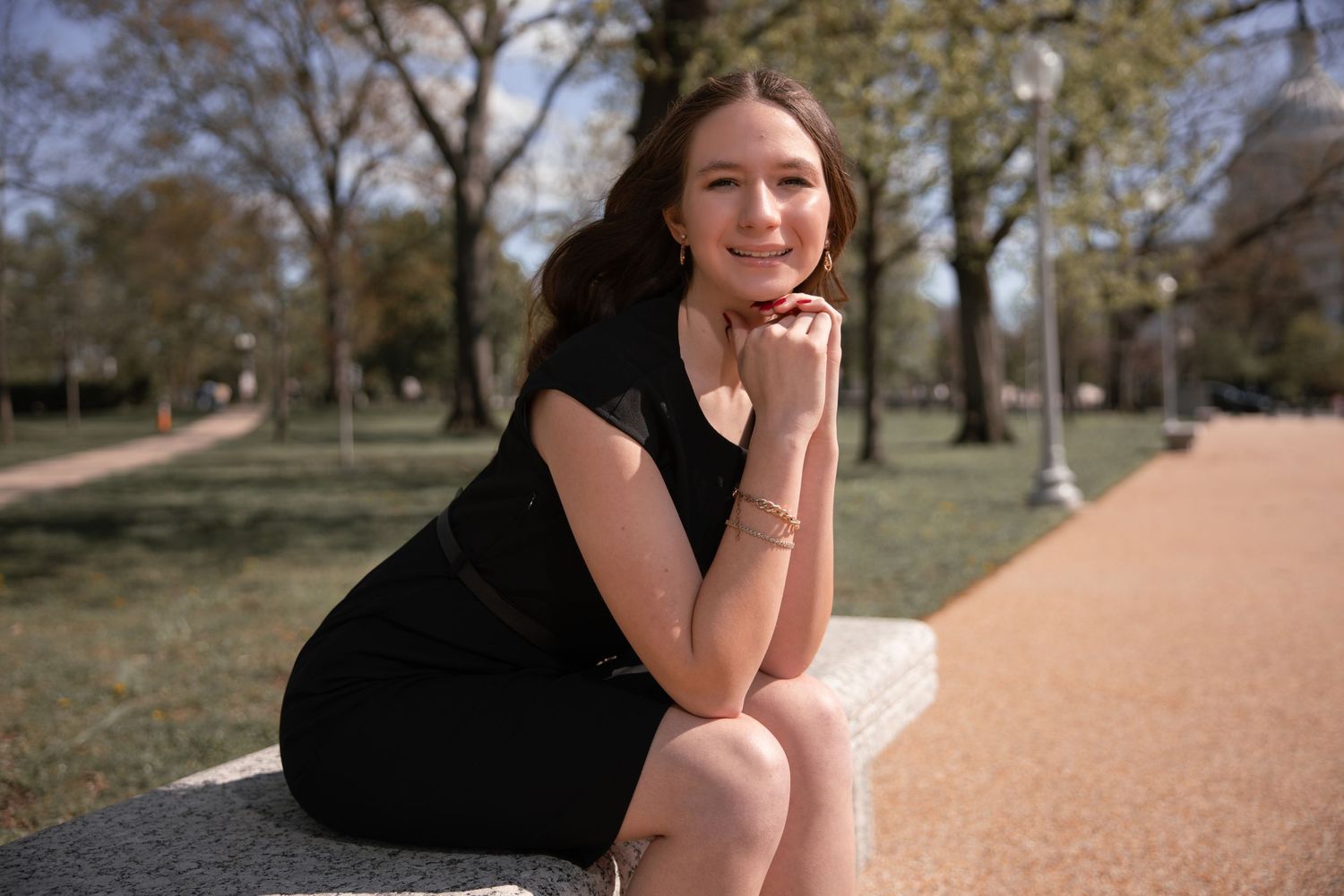 A person in a black dress sits thoughtfully on a stone wall along a winding path in a park on a sunny day.