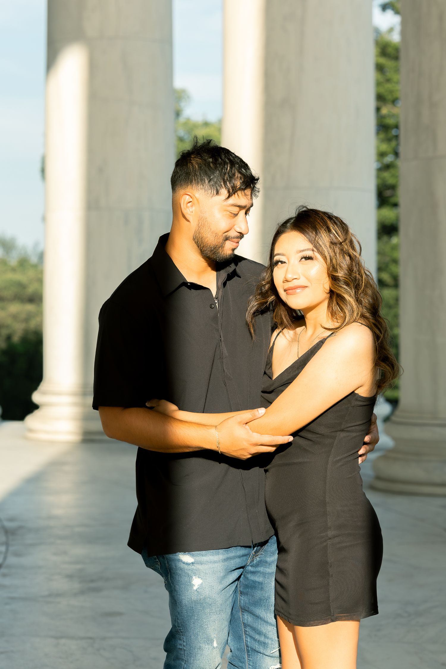 Romantic couple embraces while dancing between white columns in outdoor setting during golden hour.