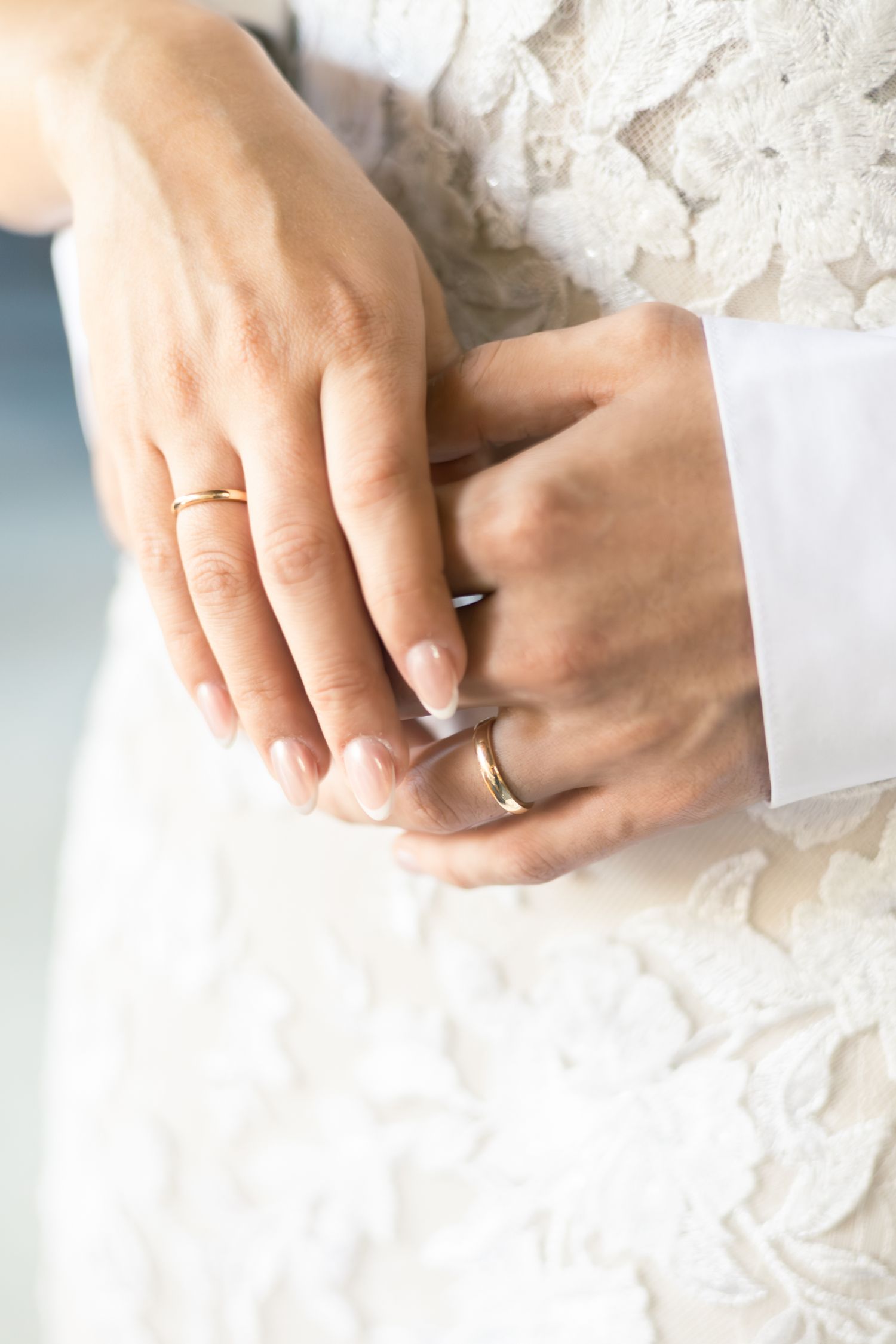 Close-up of hands with French manicure adjusting a wedding ring against white lace dress fabric.