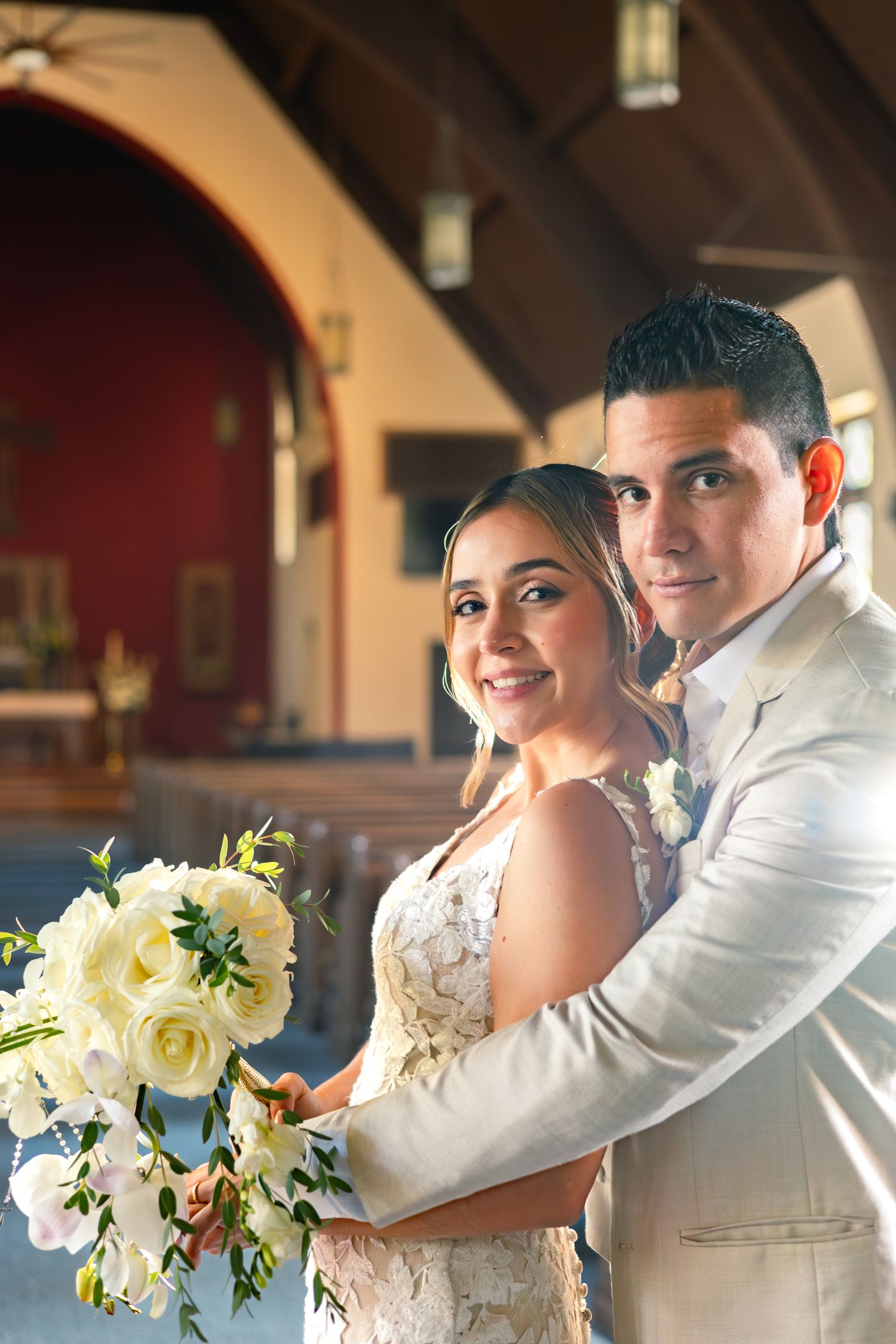A happy newlywed couple embraces in a church while holding a beautiful bouquet of white roses and flowers.